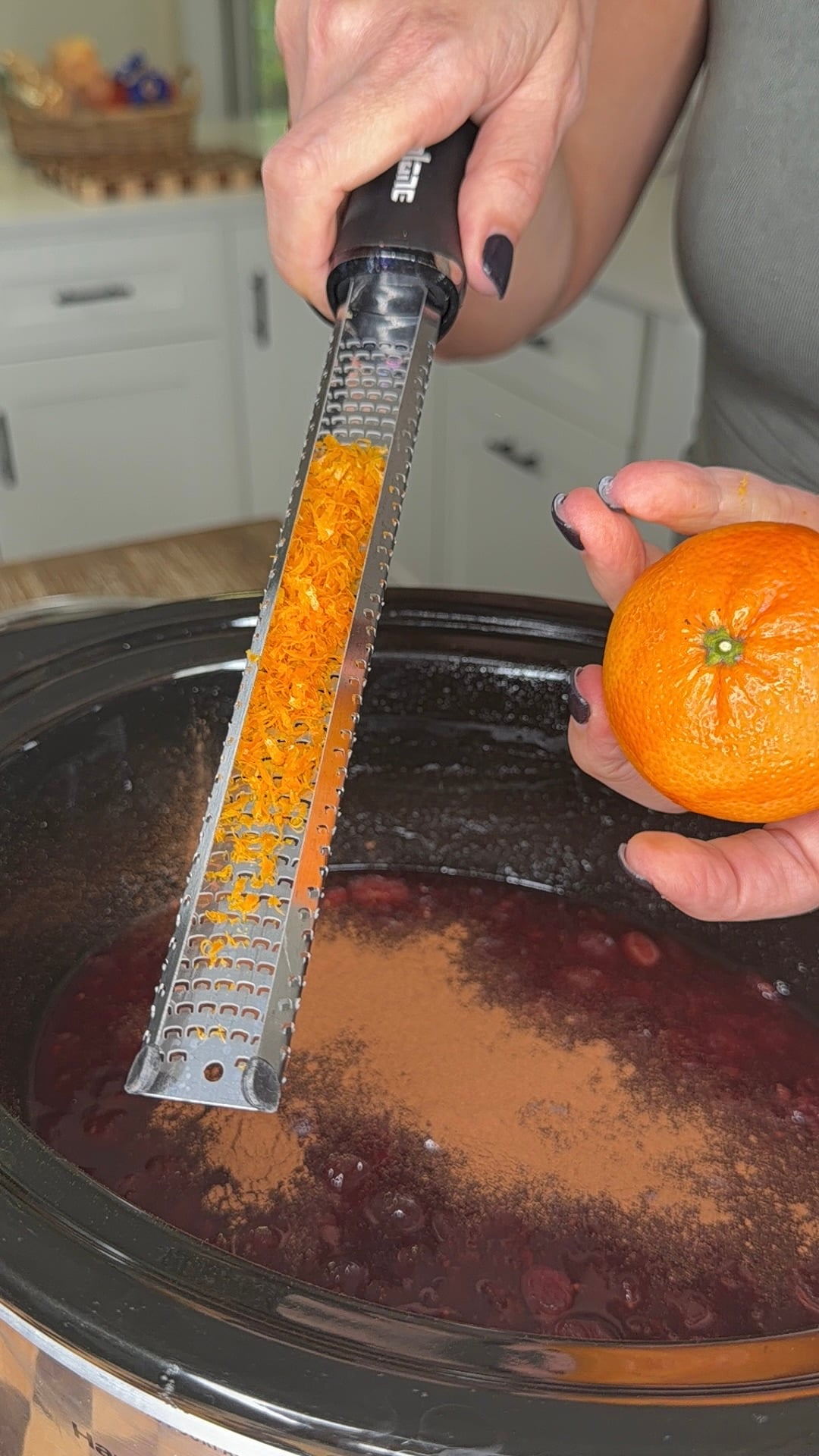 A person zesting an orange over a slow cooker filled with a dark berry mixture and a dusting of cinnamon.