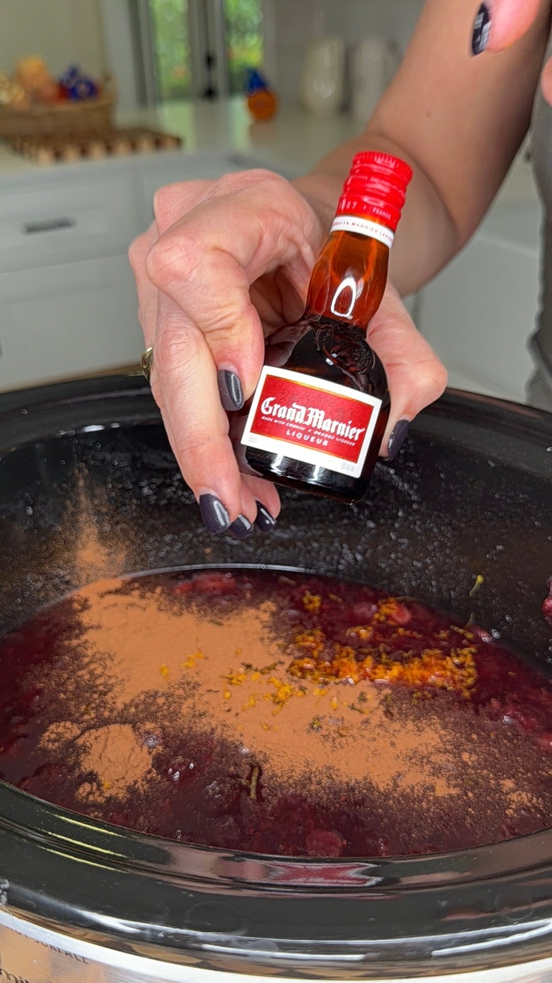 A hand pours a miniature bottle of Grand Marnier liqueur into a slow cooker filled with a dark red mixture dusted with cinnamon.