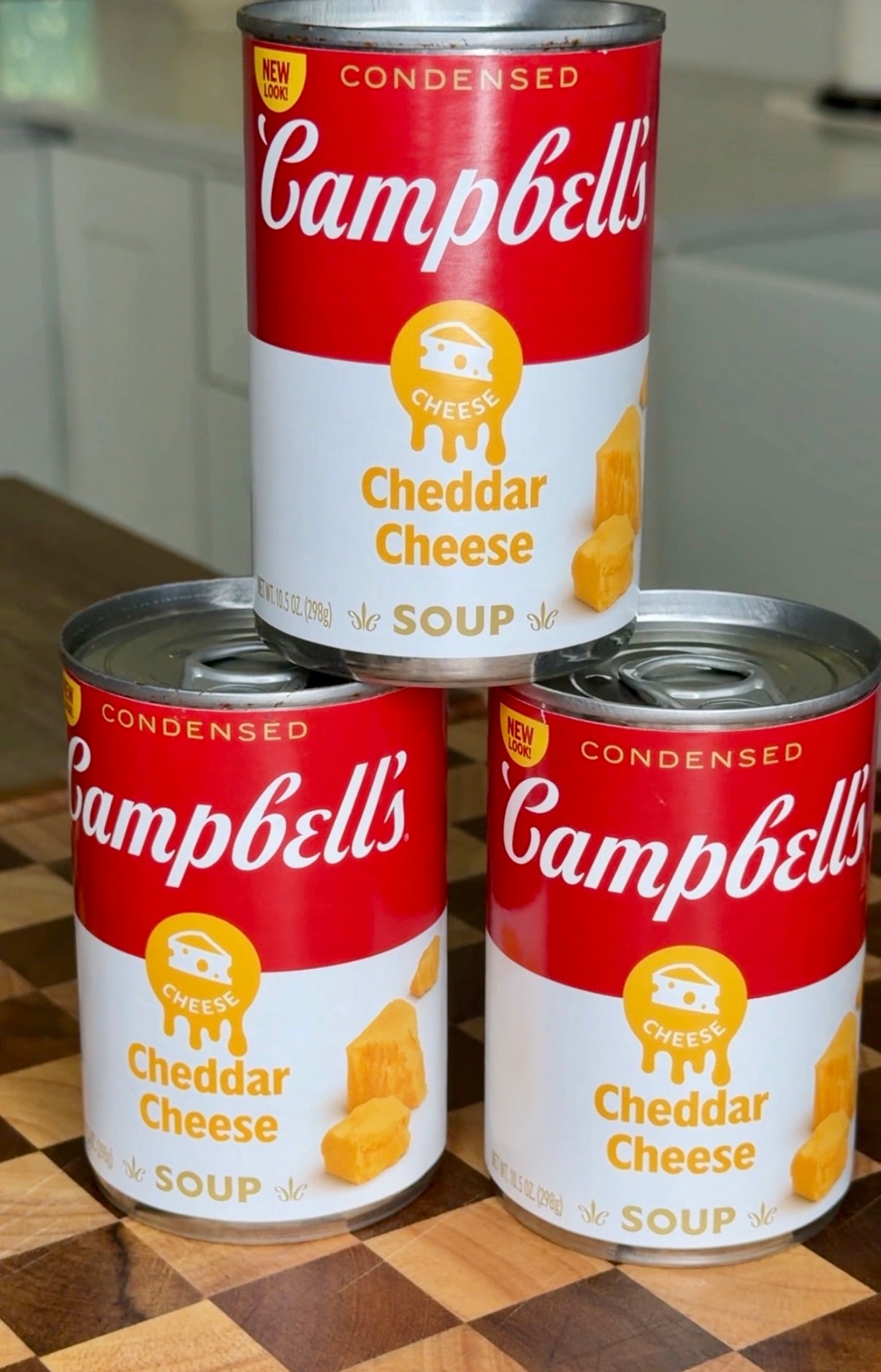 Three cans of Campbells condensed cheddar cheese soup stacked on a checkered surface.