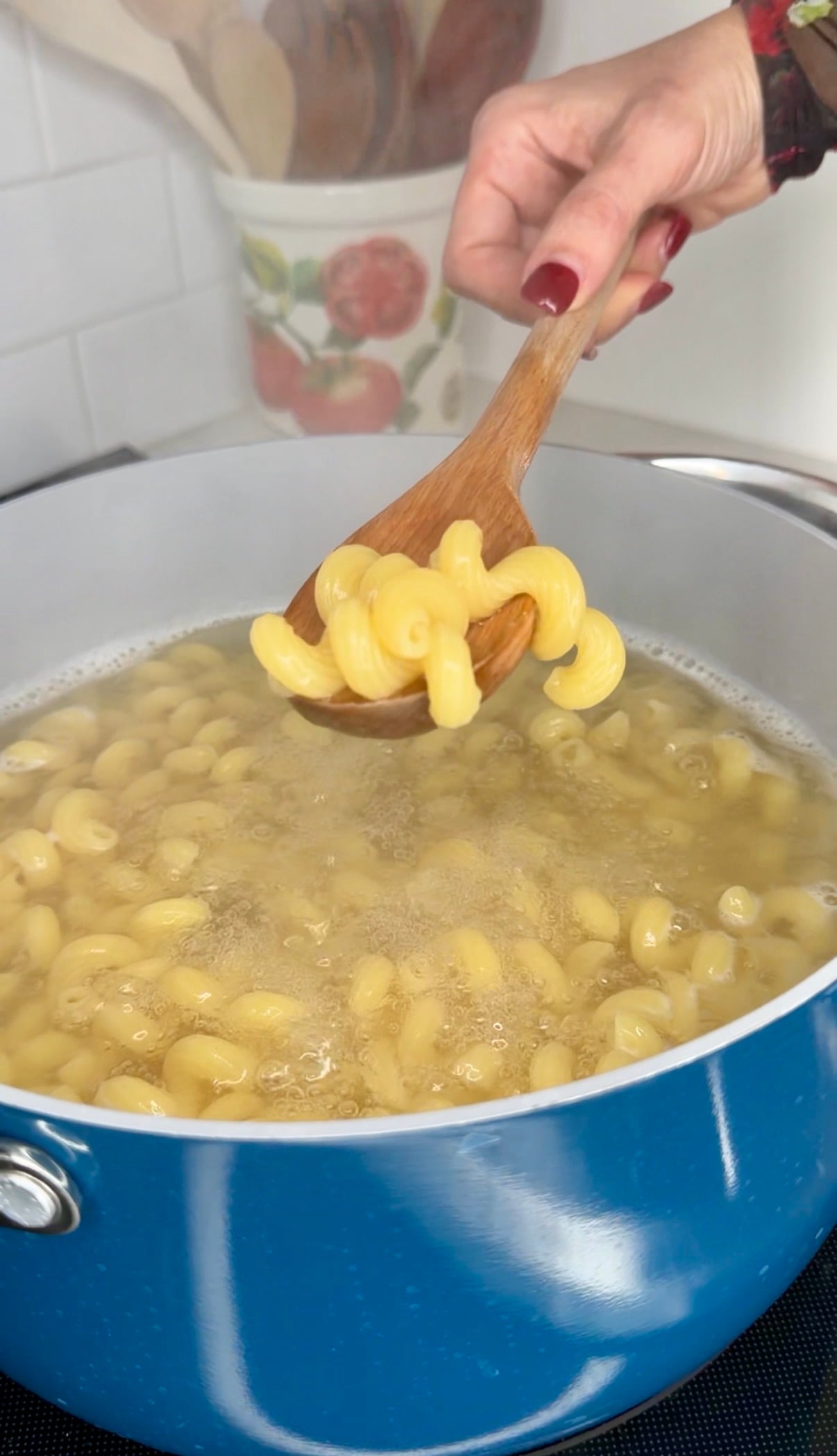 A hand holding a wooden spoon lifts cooked curly pasta from a blue pot of boiling water.