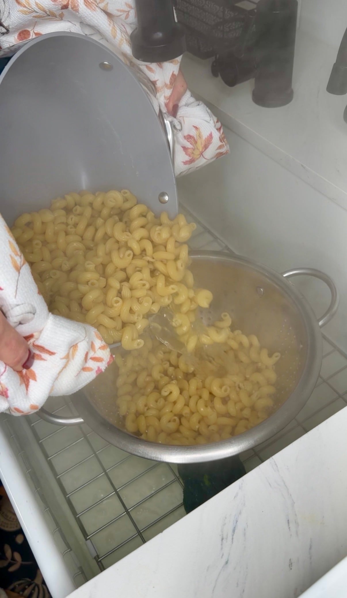 Hands wearing autumn-themed oven mitts pour cooked macaroni from a gray pot into a metal colander in a kitchen sink.
