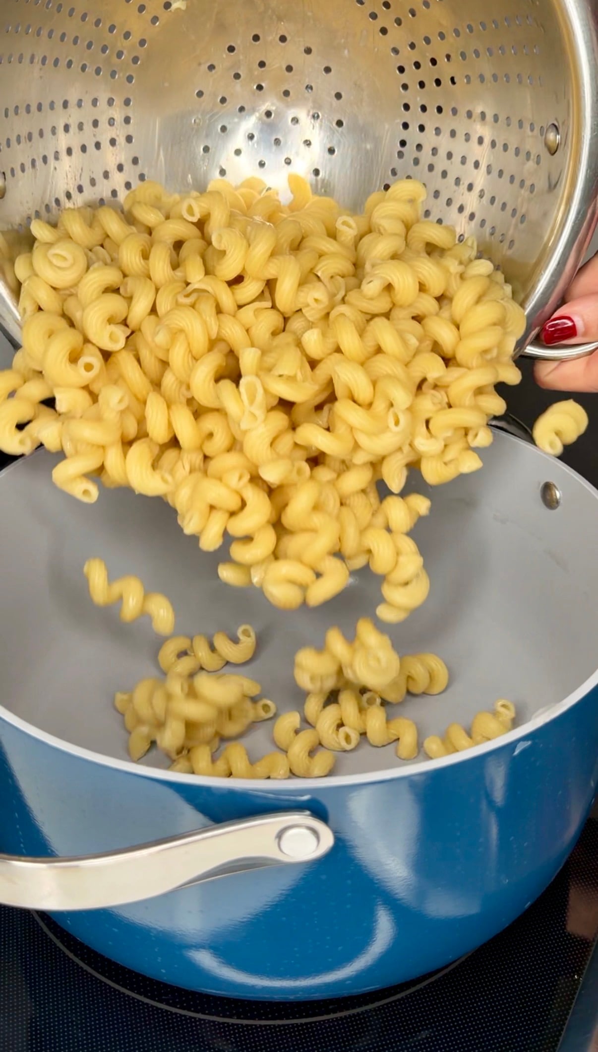 Cooked curly pasta being poured from a metal colander into a blue pot.