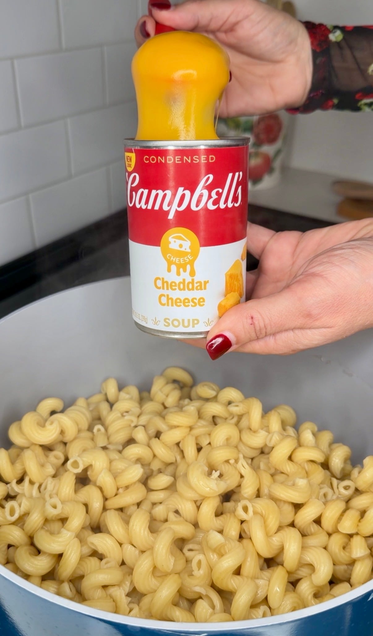 A hand squeezes a glob of yellow cheese out of an open can of Campbells Condensed Cheddar Cheese soup into a pot of cooked pasta.