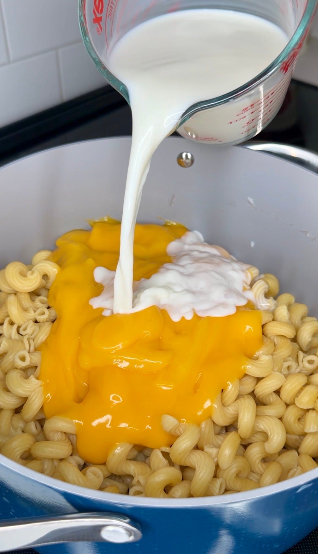 Milk being poured from a measuring cup into a pot of cooked pasta with melted cheese.