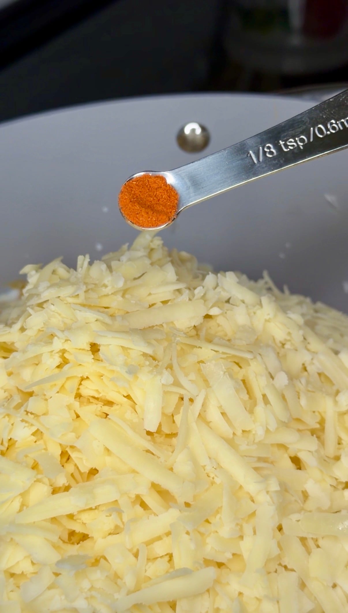 A measuring spoon holding a small amount of orange spice being poured over a pile of shredded cheese.