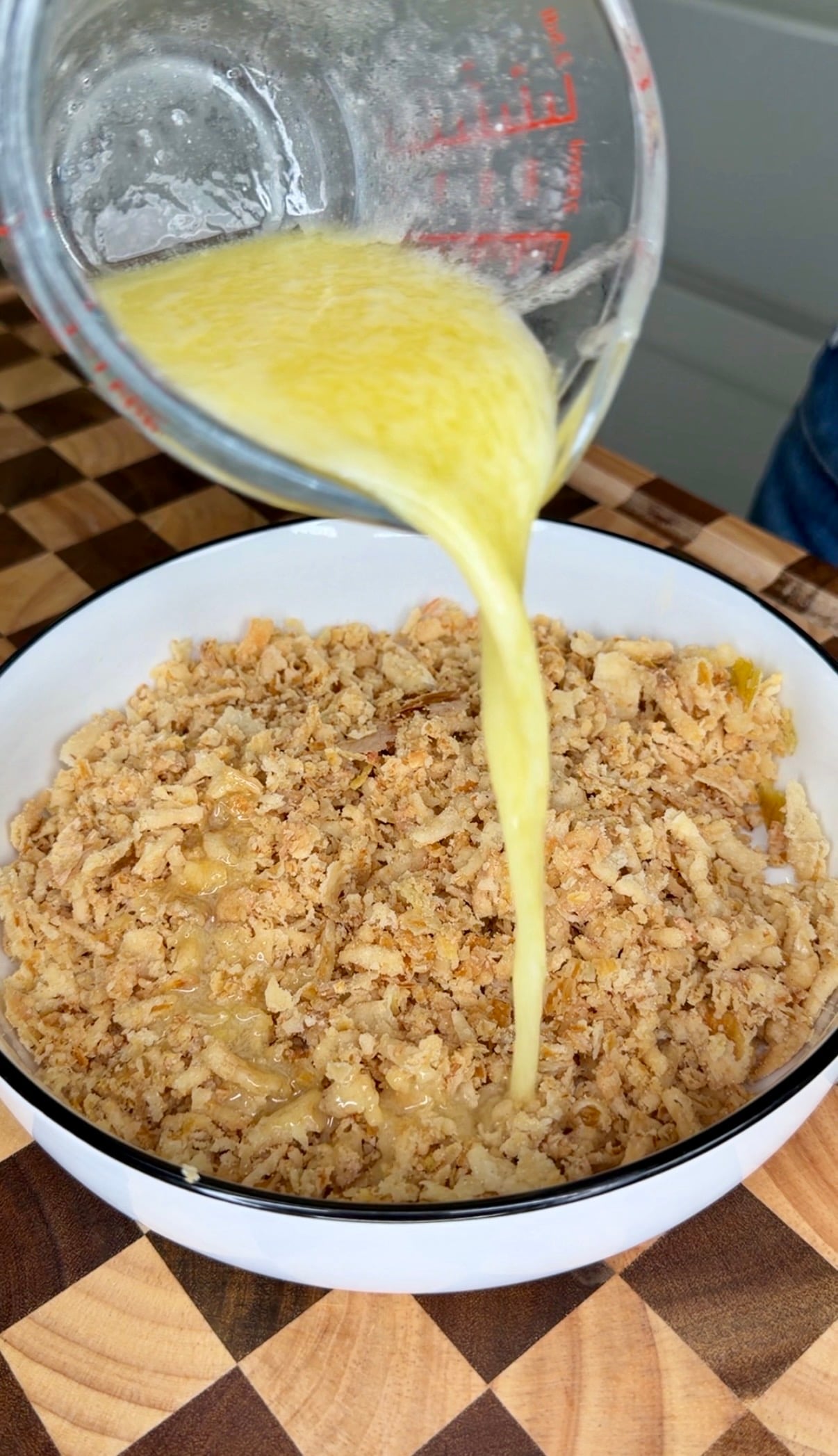 Melted butter is poured from a glass measuring cup into a white bowl filled with crushed crackers.
