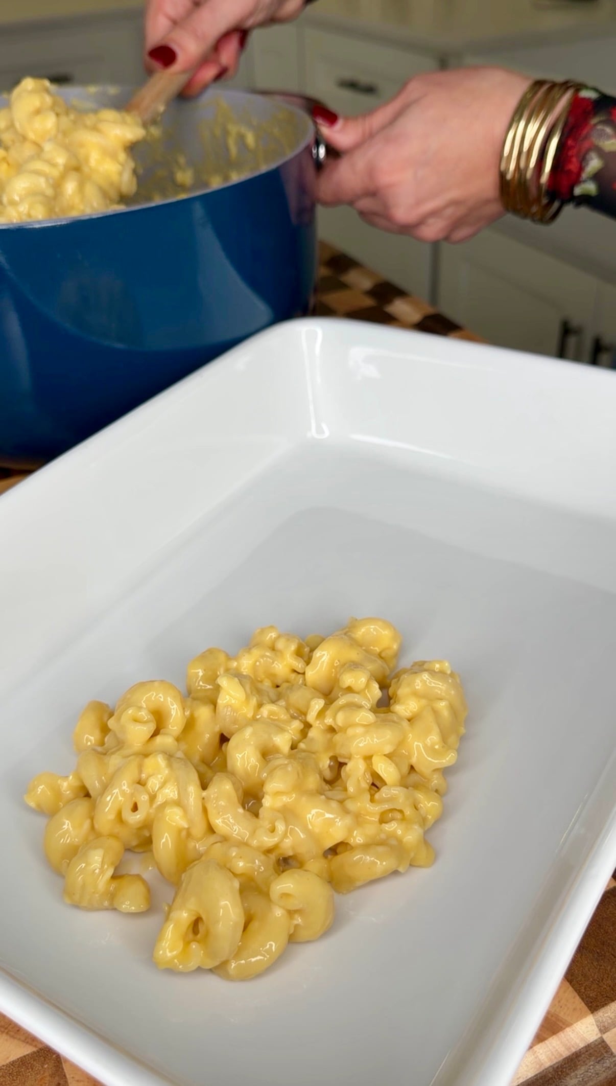 A person uses a wooden spoon to transfer macaroni and cheese from a blue pot into a white rectangular baking dish.