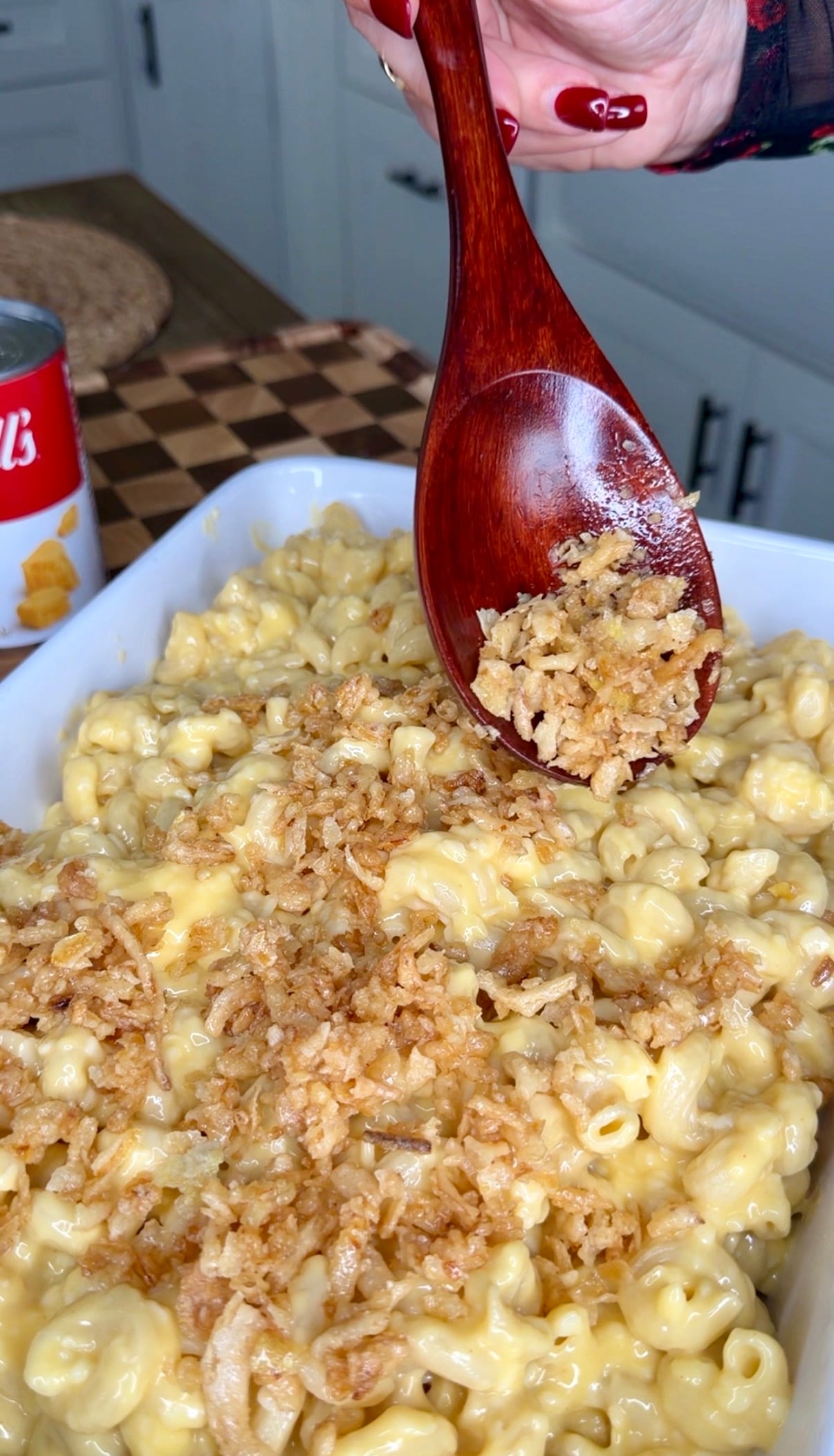 A wooden spoon lifts crispy fried onions over a baking dish of creamy macaroni and cheese.
