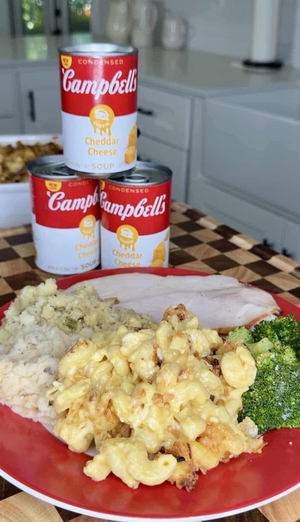 A plate with sliced turkey, macaroni and cheese, mashed potatoes, and broccoli, positioned in front of three cans of Campbells Cheddar Cheese soup.