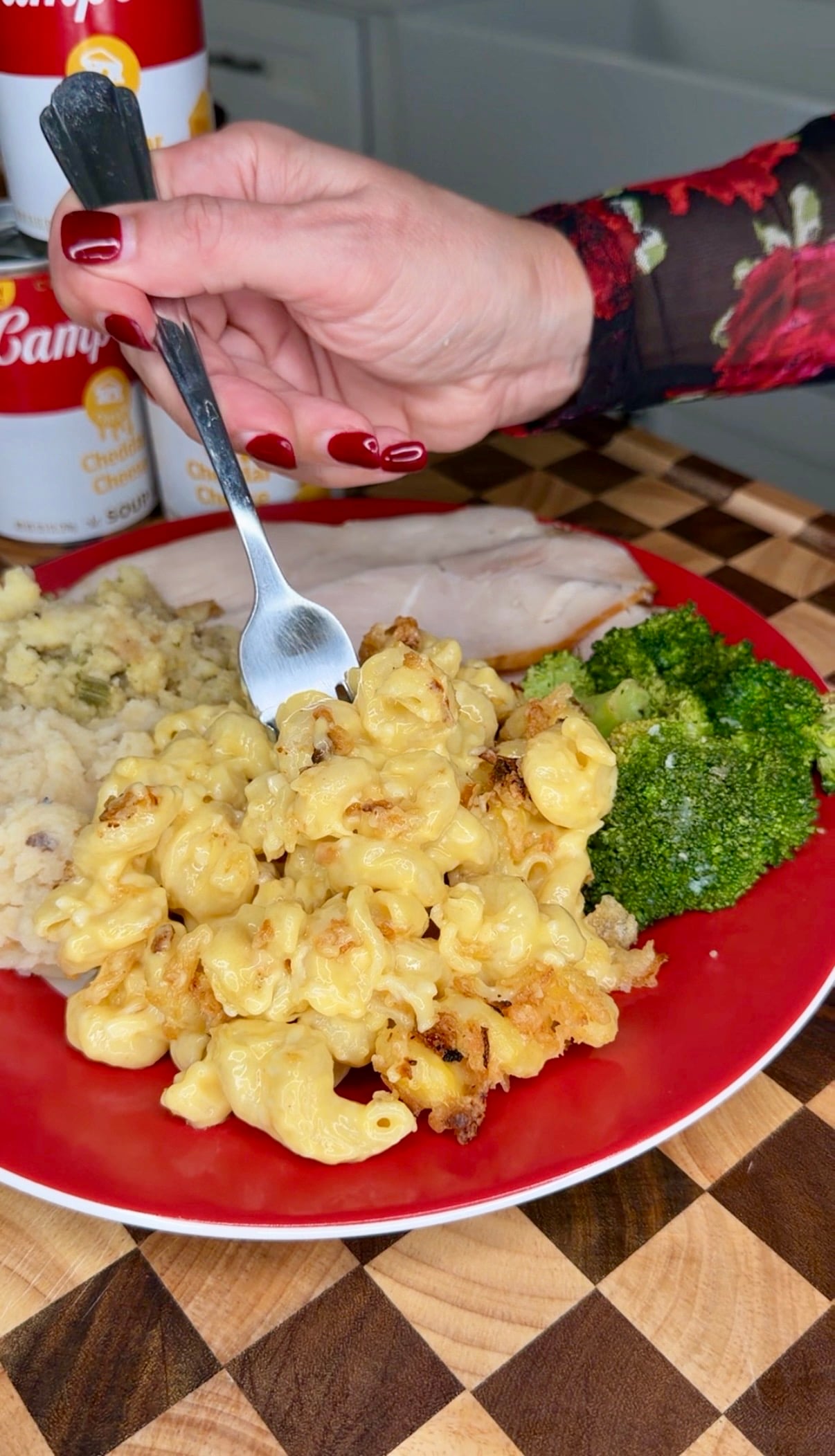 A hand holds a forkful of baked macaroni and cheese from a red plate that also contains sliced turkey, mashed potatoes, and broccoli.