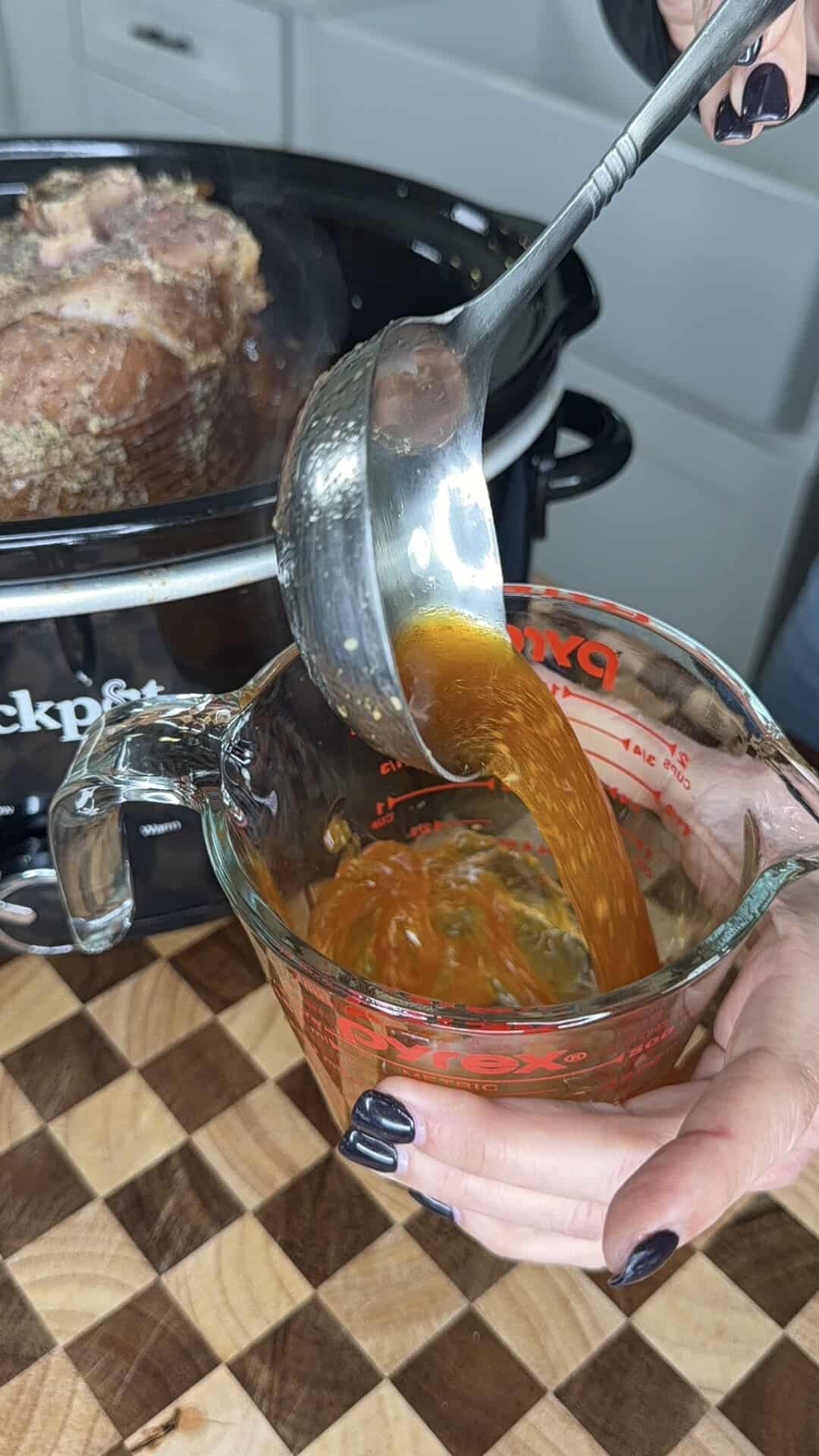 A metal ladle pours liquid drippings from a slow cooker into a glass measuring cup.