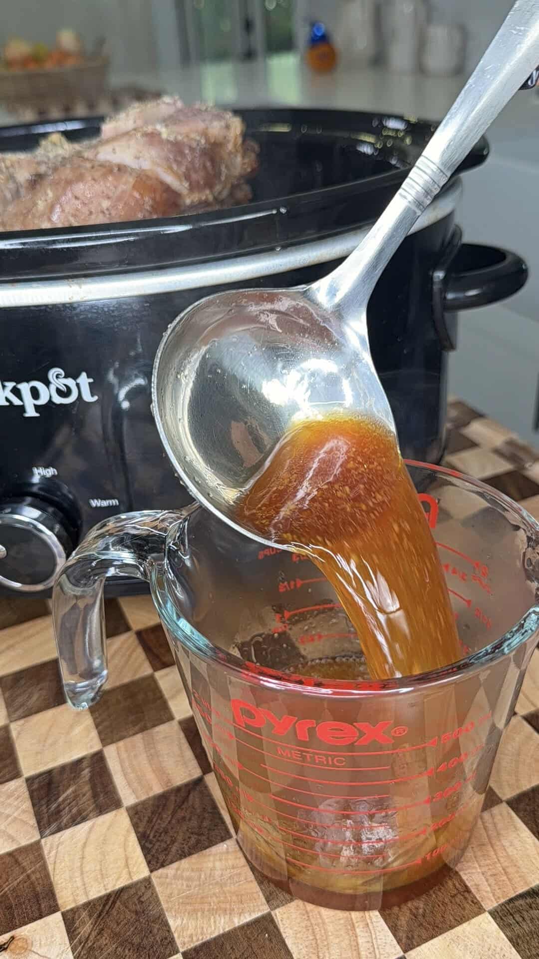 A ladle pours brown liquid from a slow cooker into a glass measuring cup.