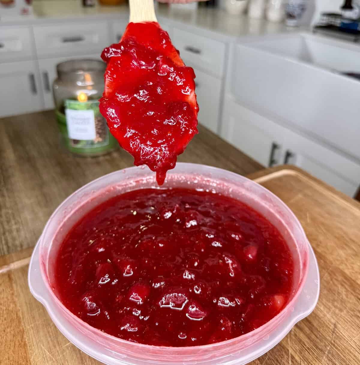 A hand holding a wooden spoon dripping with bright red cranberry sauce over a plastic container filled with the same sauce.