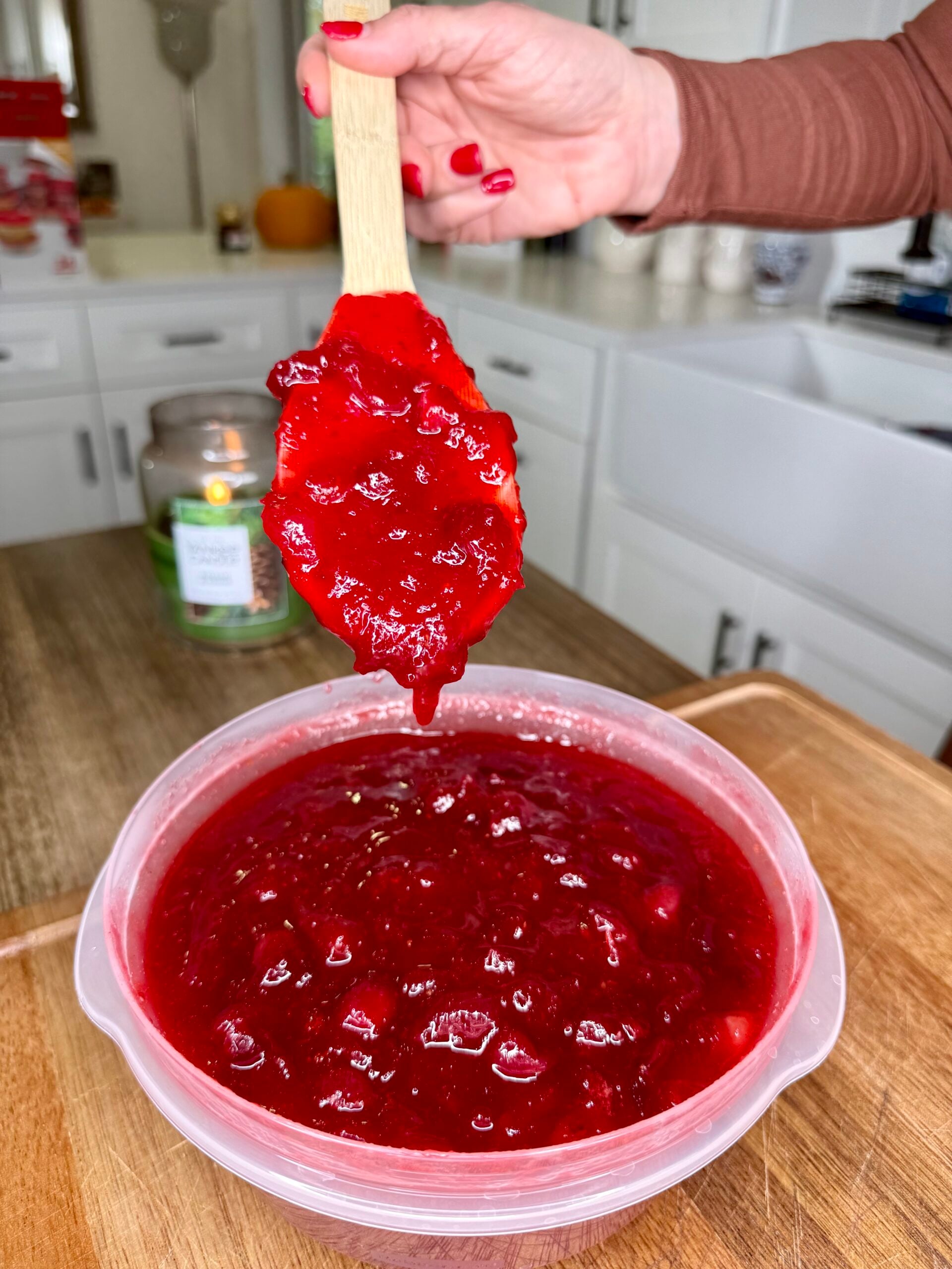 A hand holds a wooden spoon dripping with bright red cranberry sauce over a bowl filled with the same sauce.