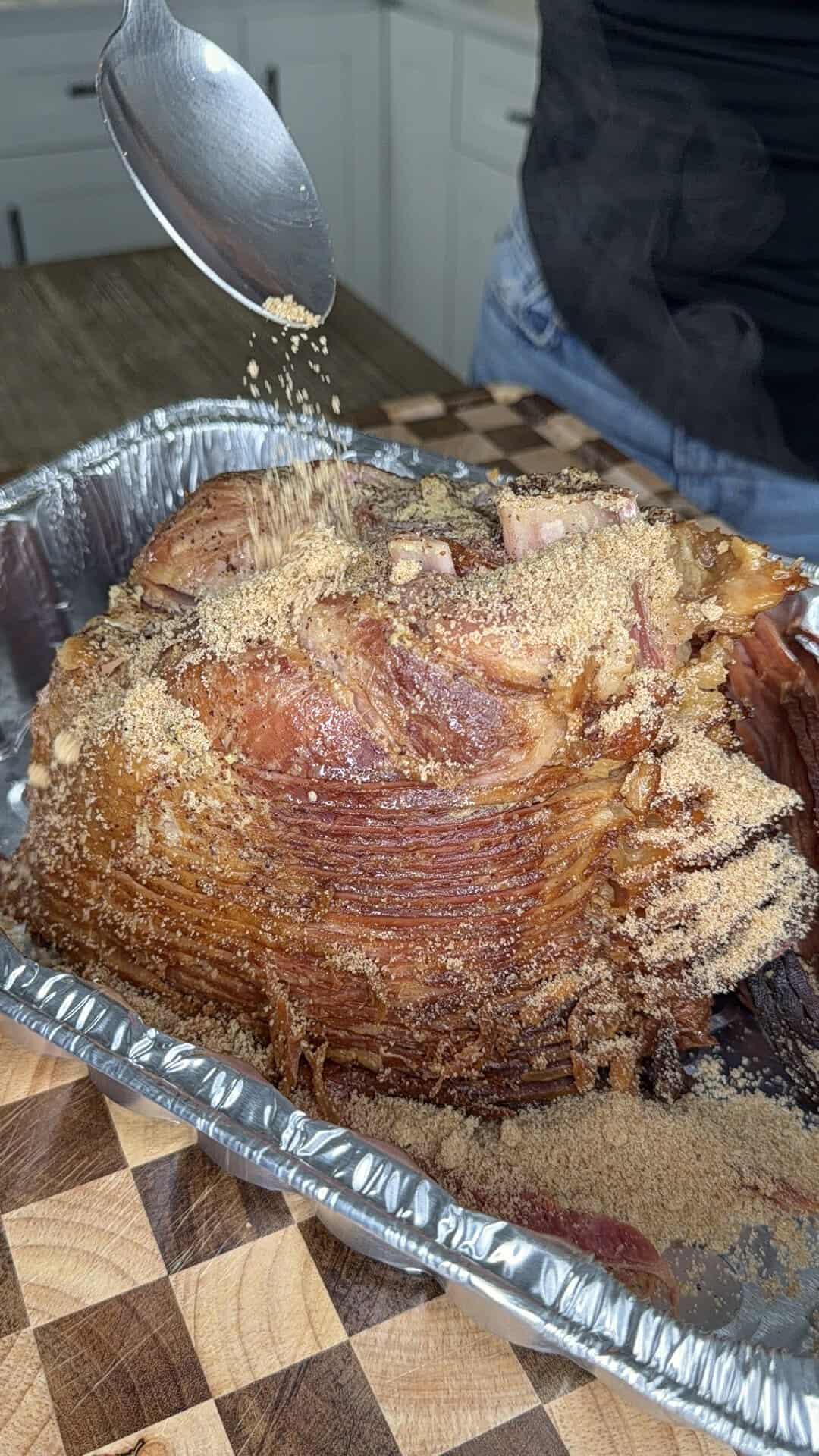 A person sprinkles brown sugar from a spoon onto a spiral-cut ham in a foil pan.