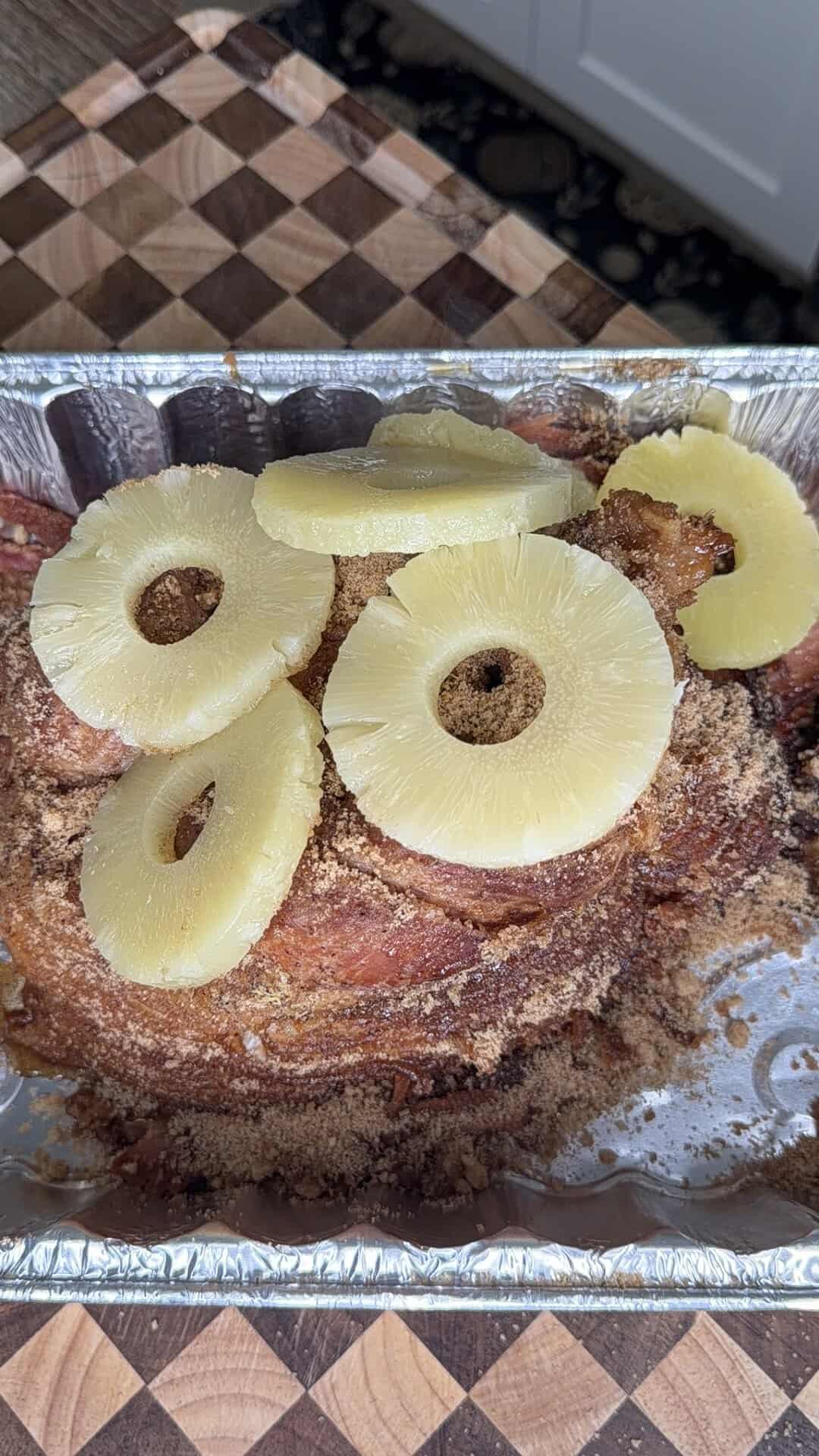 A spiral-sliced ham topped with brown sugar and pineapple rings in a foil roasting pan.