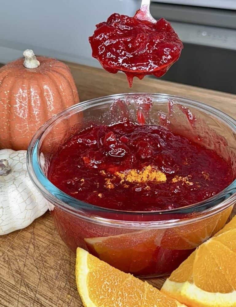 A spoonful of cranberry sauce held over a glass bowl of the same sauce, garnished with orange zest and served alongside orange slices and small pumpkins.