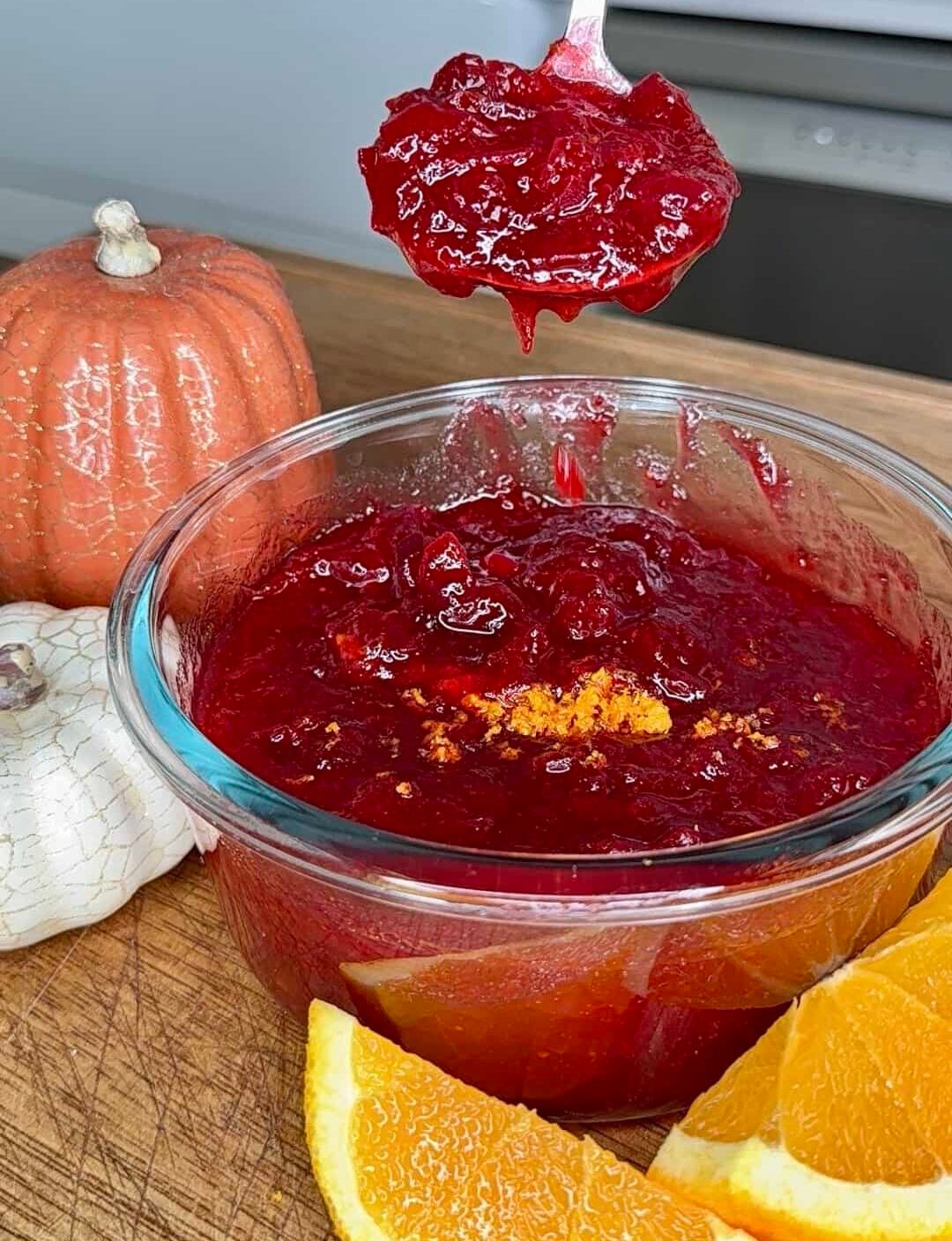 A spoonful of cranberry sauce held over a glass bowl of the same sauce, garnished with orange zest and served alongside orange slices and small pumpkins.