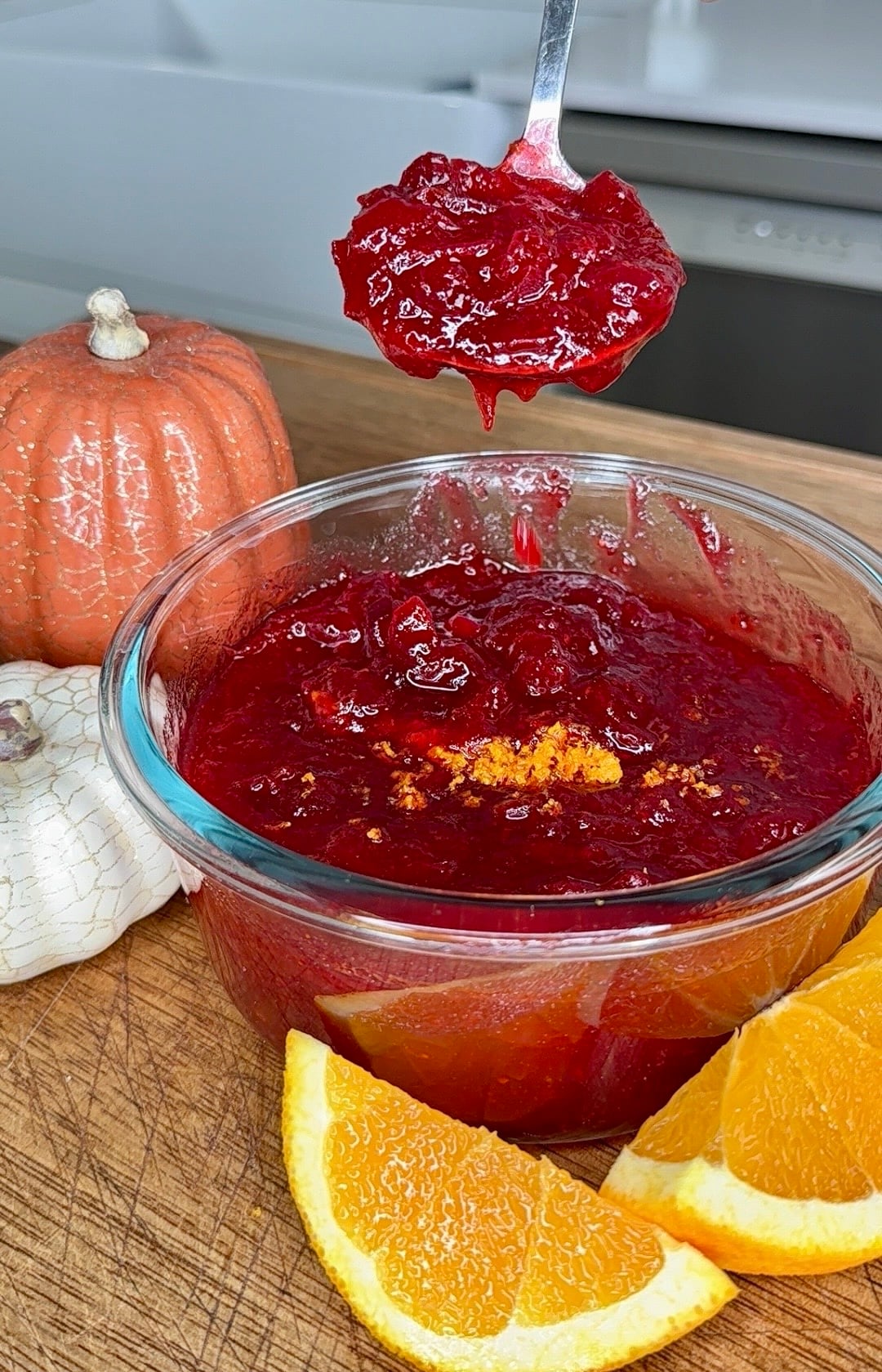 A spoonful of cranberry sauce held over a glass bowl of the same sauce, garnished with orange zest and served alongside orange slices and small pumpkins.
