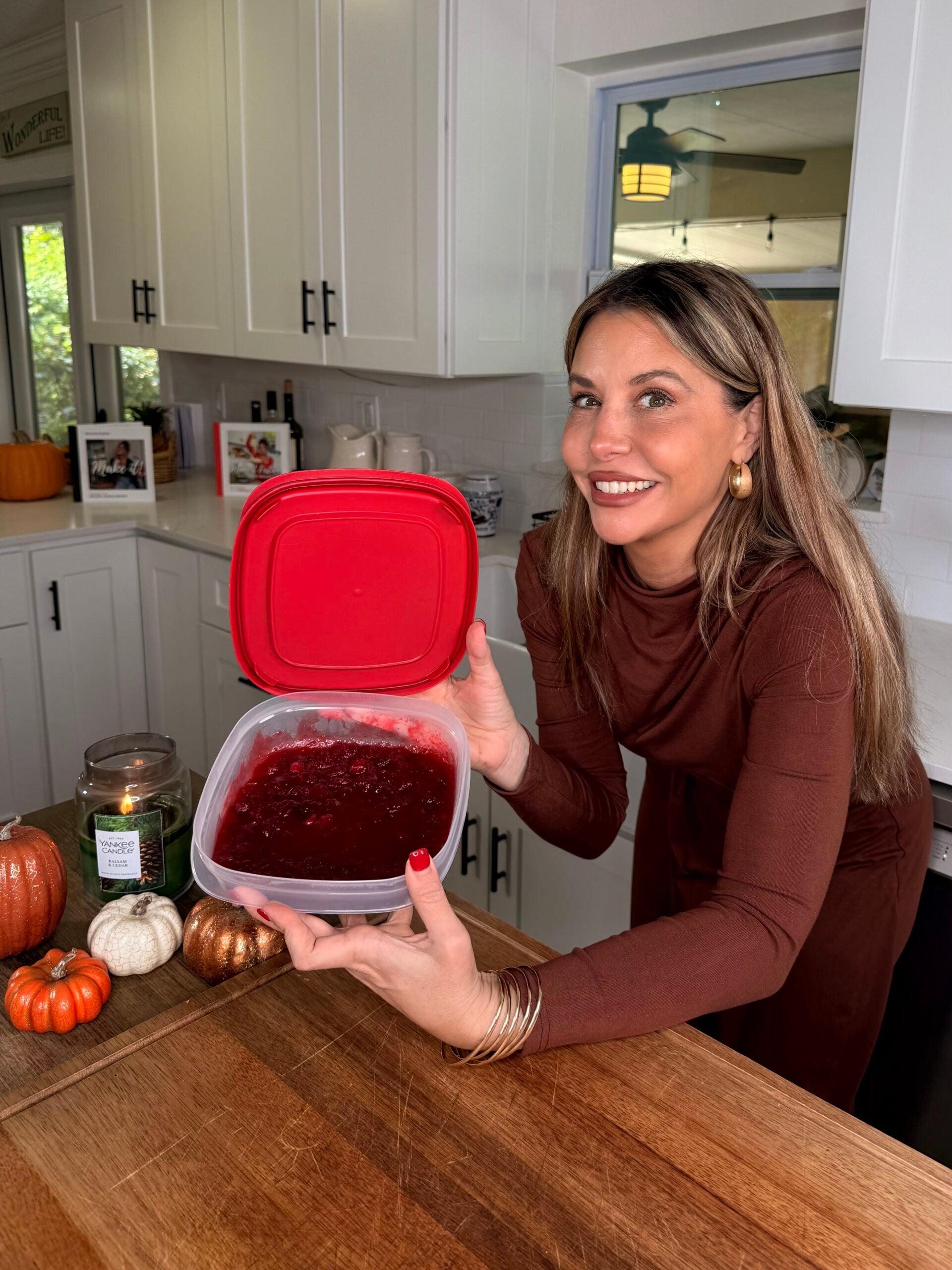 A woman smiling in a kitchen while holding a plastic container filled with cranberry sauce.