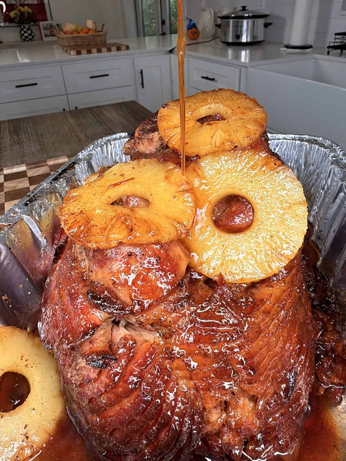 A glazed holiday ham in a foil pan, topped with pineapple rings and being drizzled with syrup.