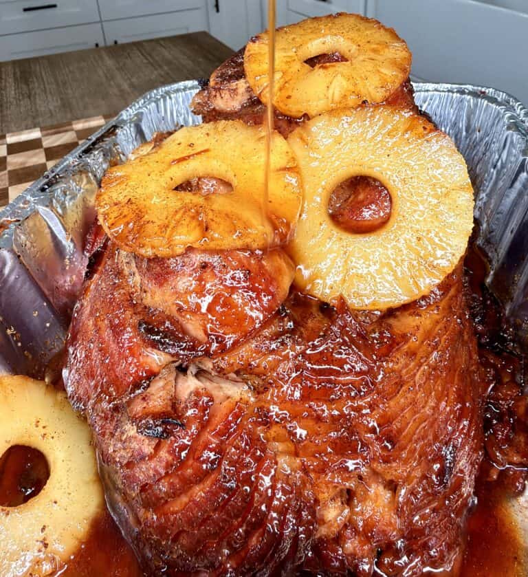 Glazed spiral-cut ham topped with pineapple rings, with syrup being poured over the top in a metal roasting pan.