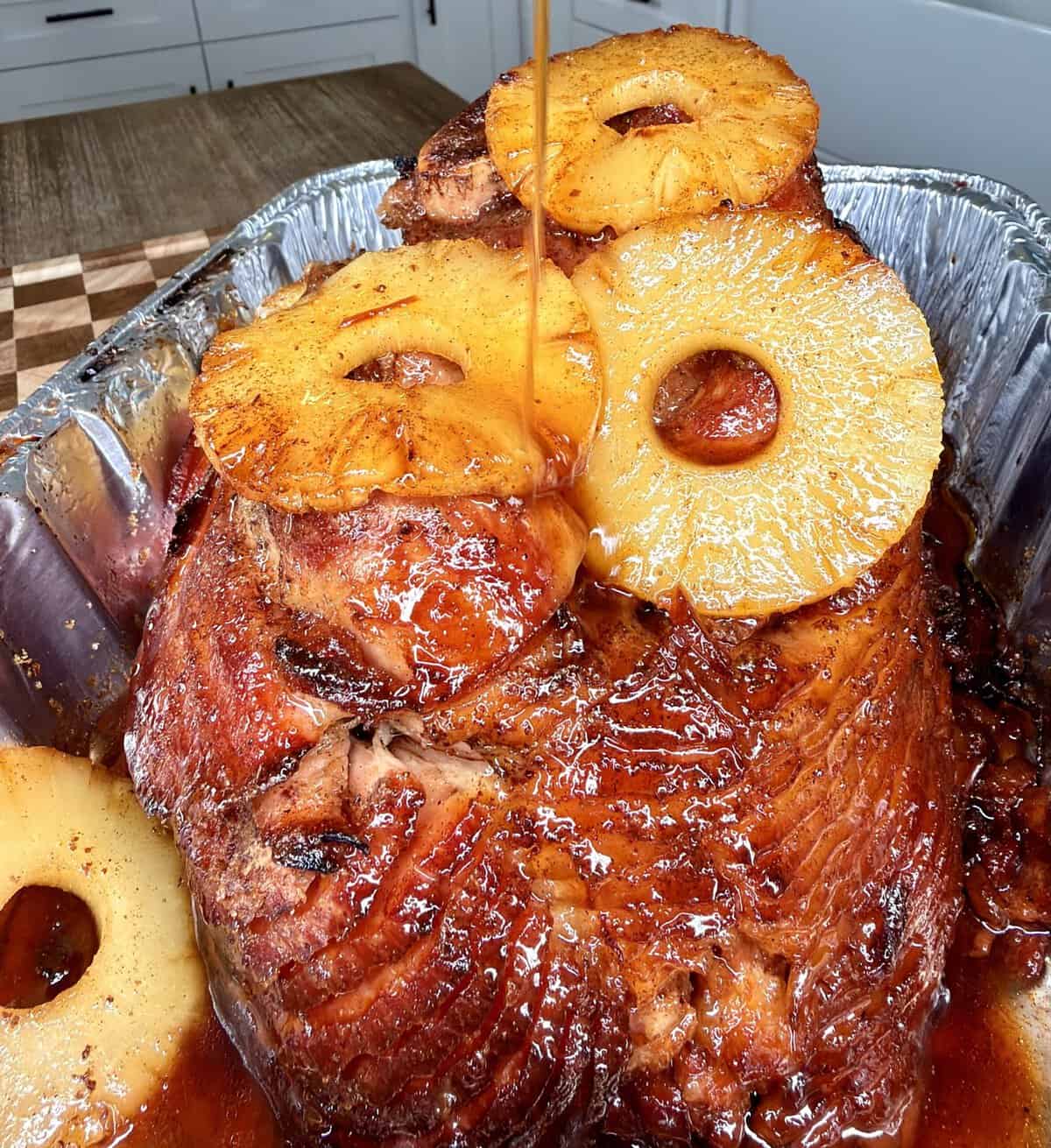 Glazed spiral-cut ham topped with pineapple rings, with syrup being poured over the top in a metal roasting pan.