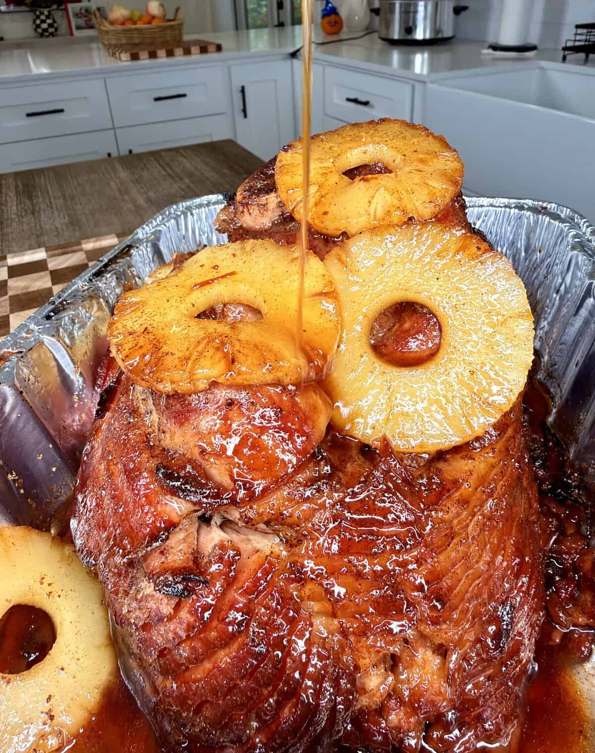 Glazed spiral-cut ham topped with pineapple rings as syrup is poured over it in a foil roasting pan.