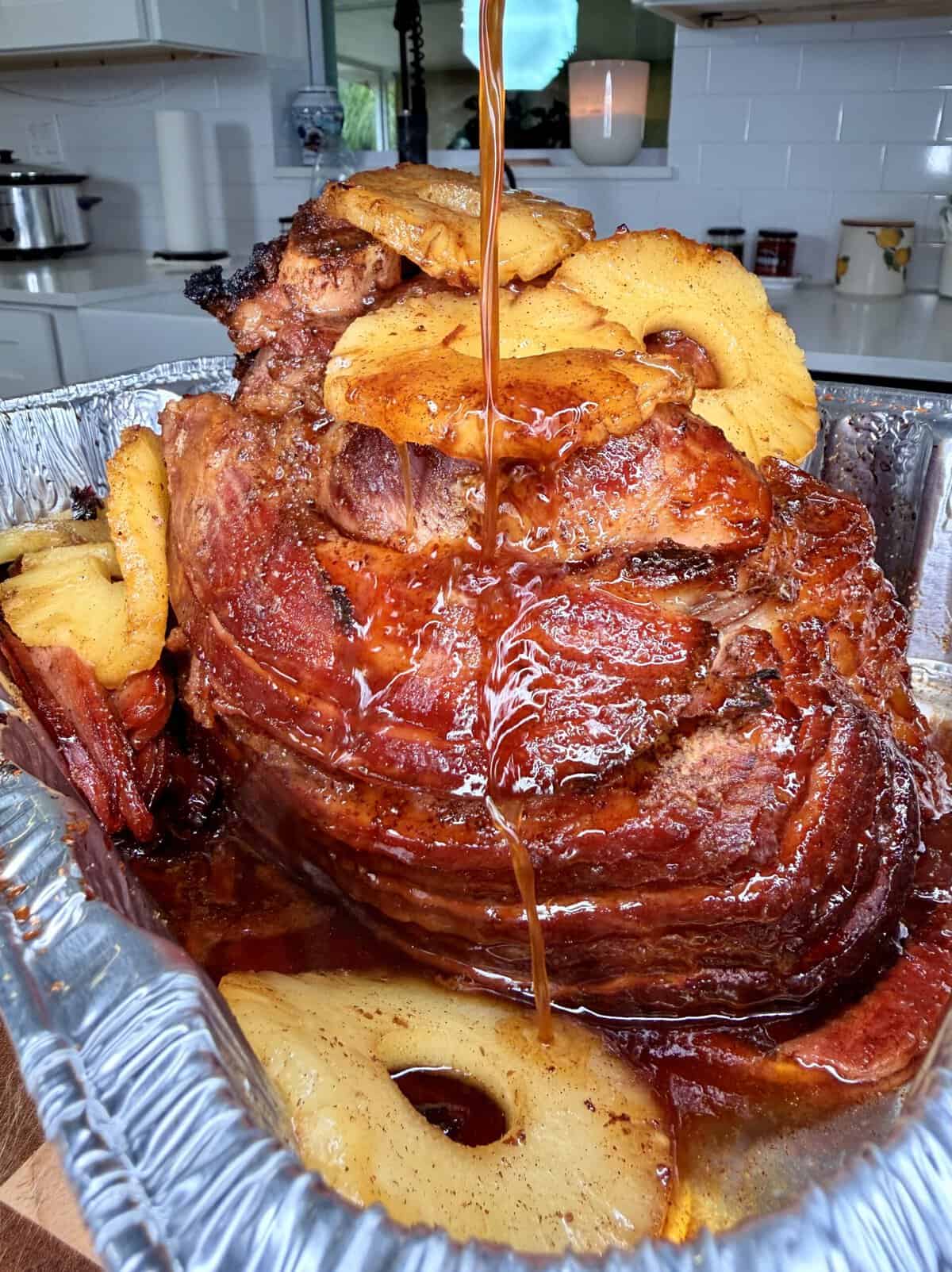 Glaze being poured over a spiral-cut ham topped with pineapple rings in a foil roasting pan.