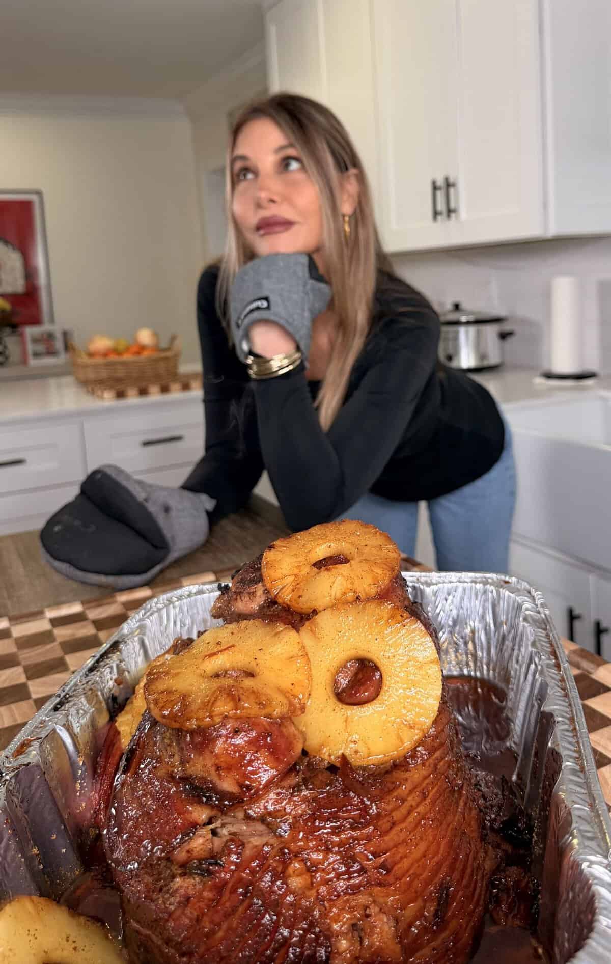 A woman in an oven mitt rests her chin on her hand, looking toward a glazed ham topped with pineapple rings in a roasting pan.