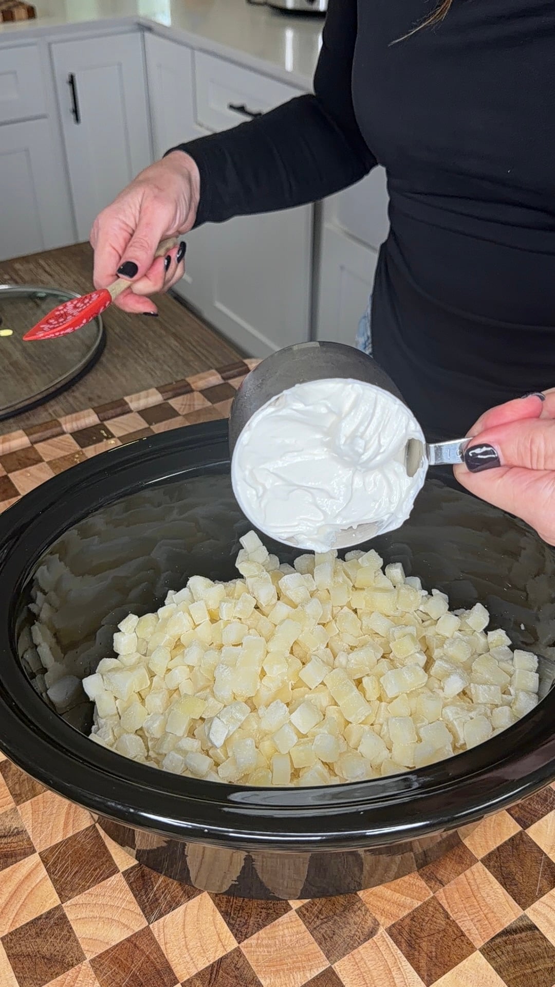 A person pouring a measuring cup of sour cream into a slow cooker filled with diced potatoes.