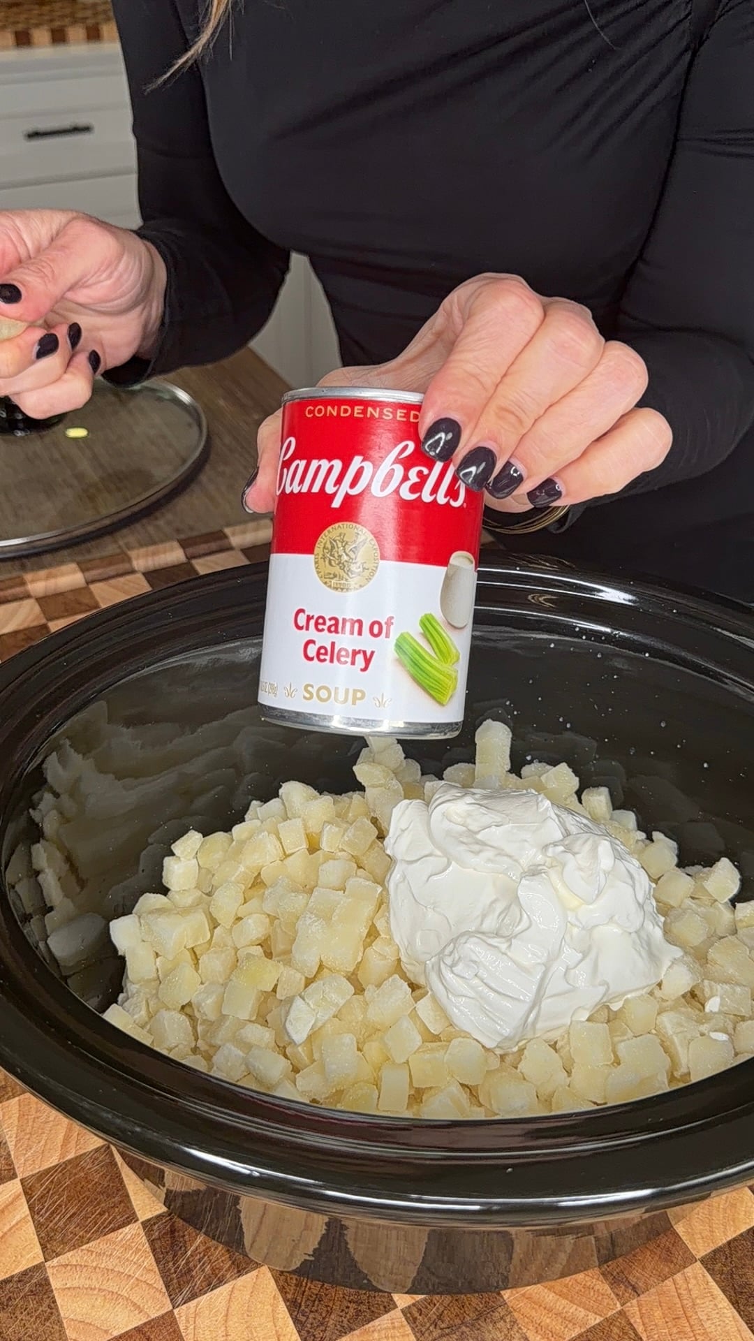 A person holds a can of Campbells Cream of Celery soup over a slow cooker filled with diced potatoes and a dollop of sour cream.