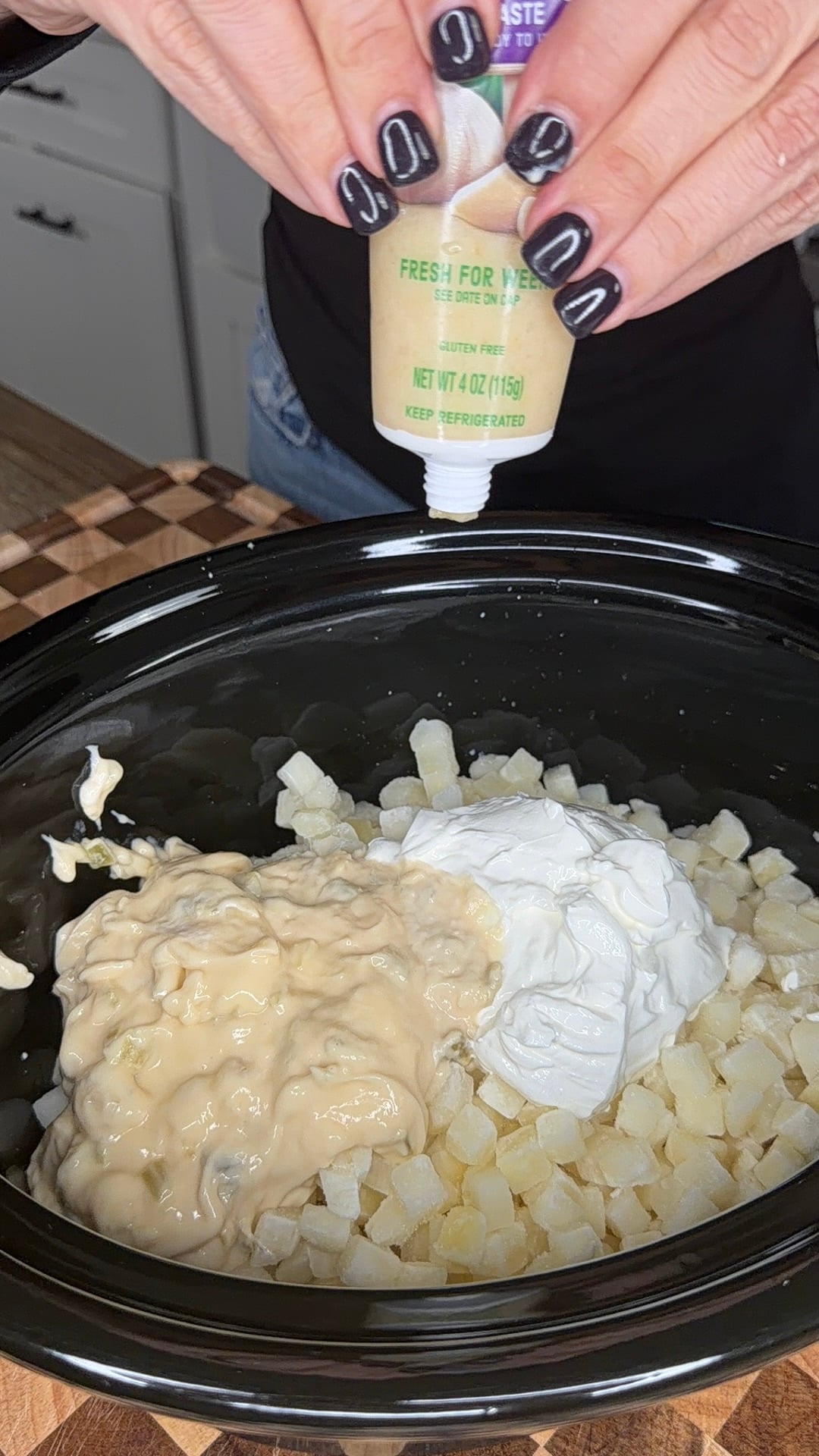 Hands squeezing a tube of garlic paste into a slow cooker filled with diced potatoes, sour cream, and a creamy sauce.