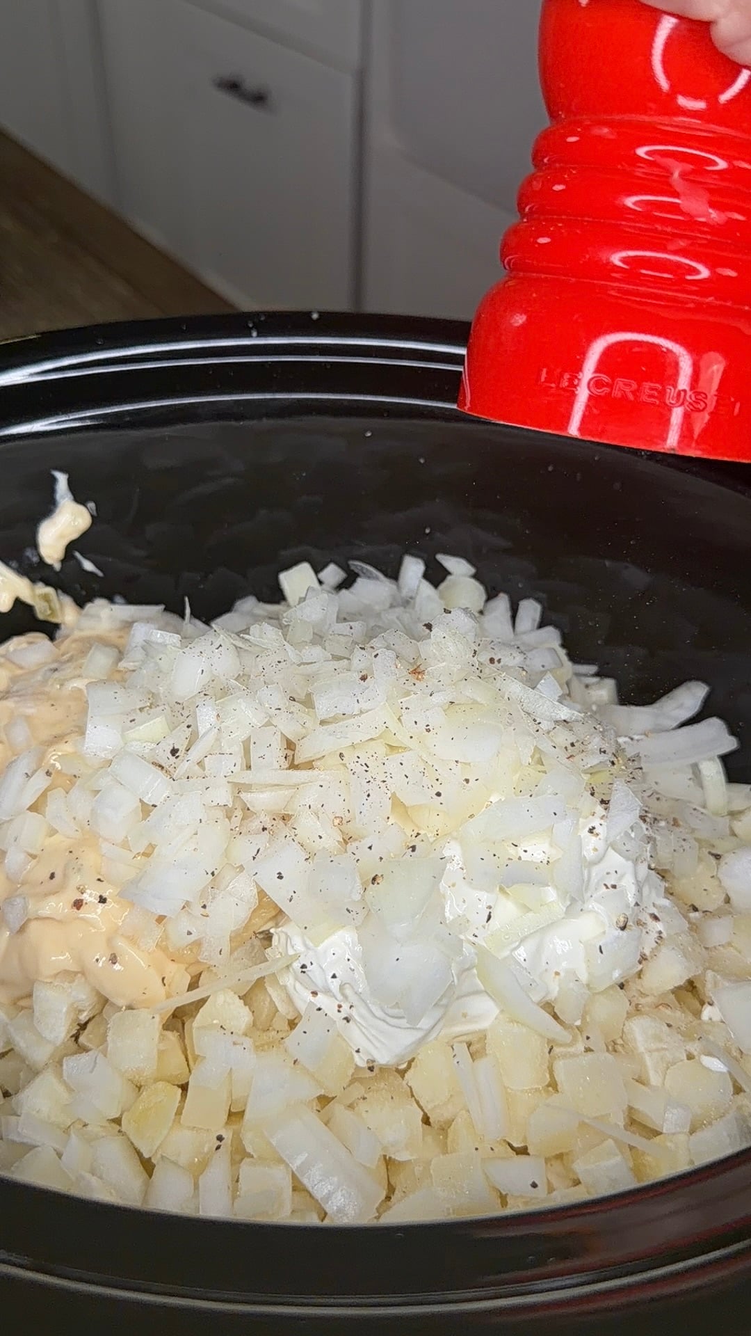 A red salt or pepper mill seasoning a slow cooker filled with diced onions, mayonnaise, and other ingredients.