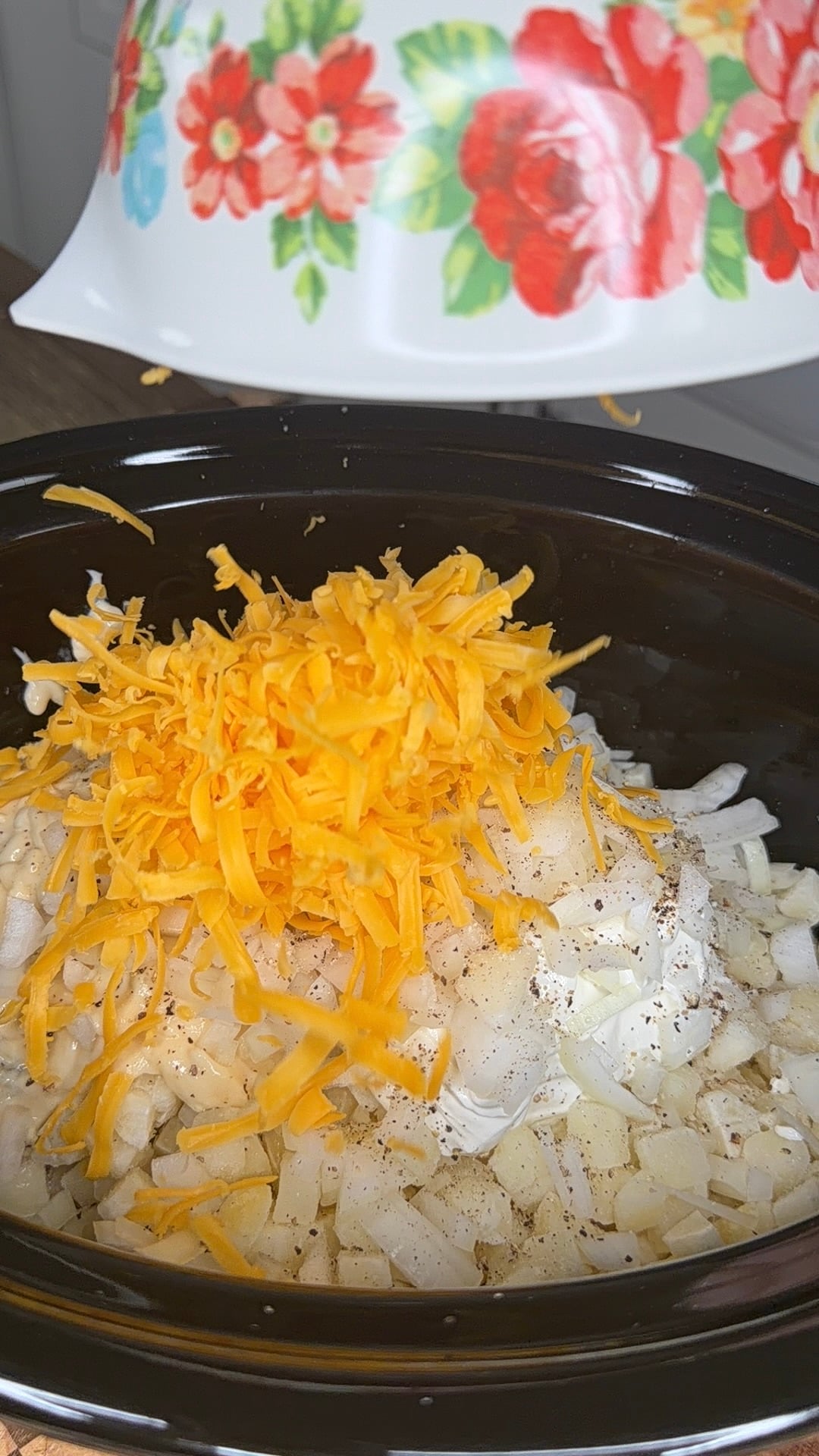 Shredded cheddar cheese being poured from a floral bowl into a slow cooker containing diced onions, cream, and seasonings.