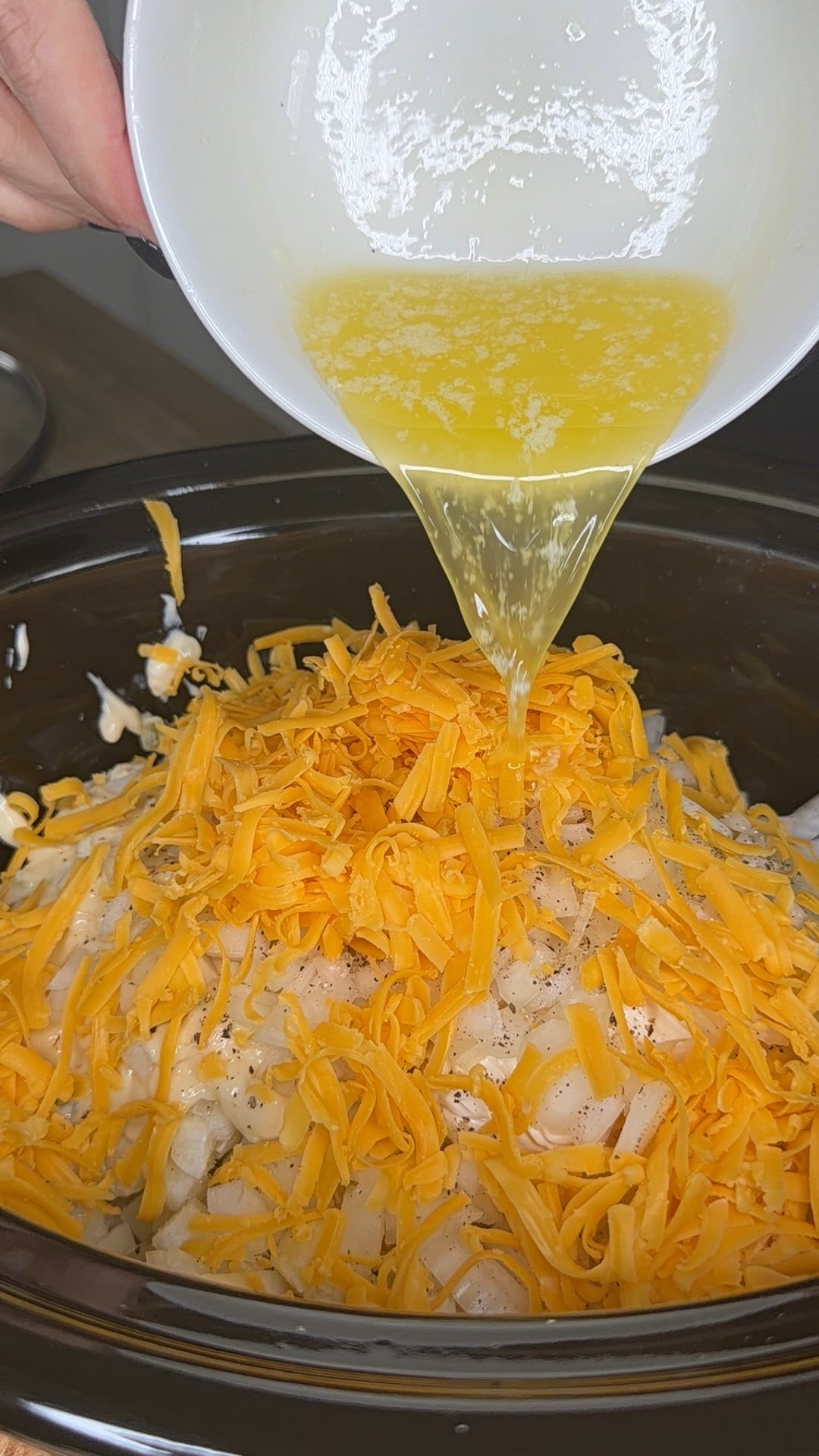 Melted butter being poured from a white bowl over a mound of shredded cheddar cheese and diced onions in a slow cooker.