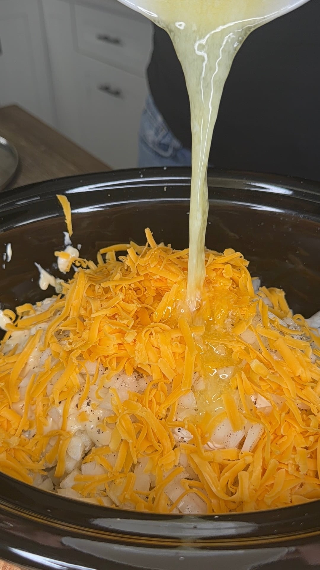 Liquid being poured from a bowl into a slow cooker filled with shredded cheddar cheese and other ingredients.