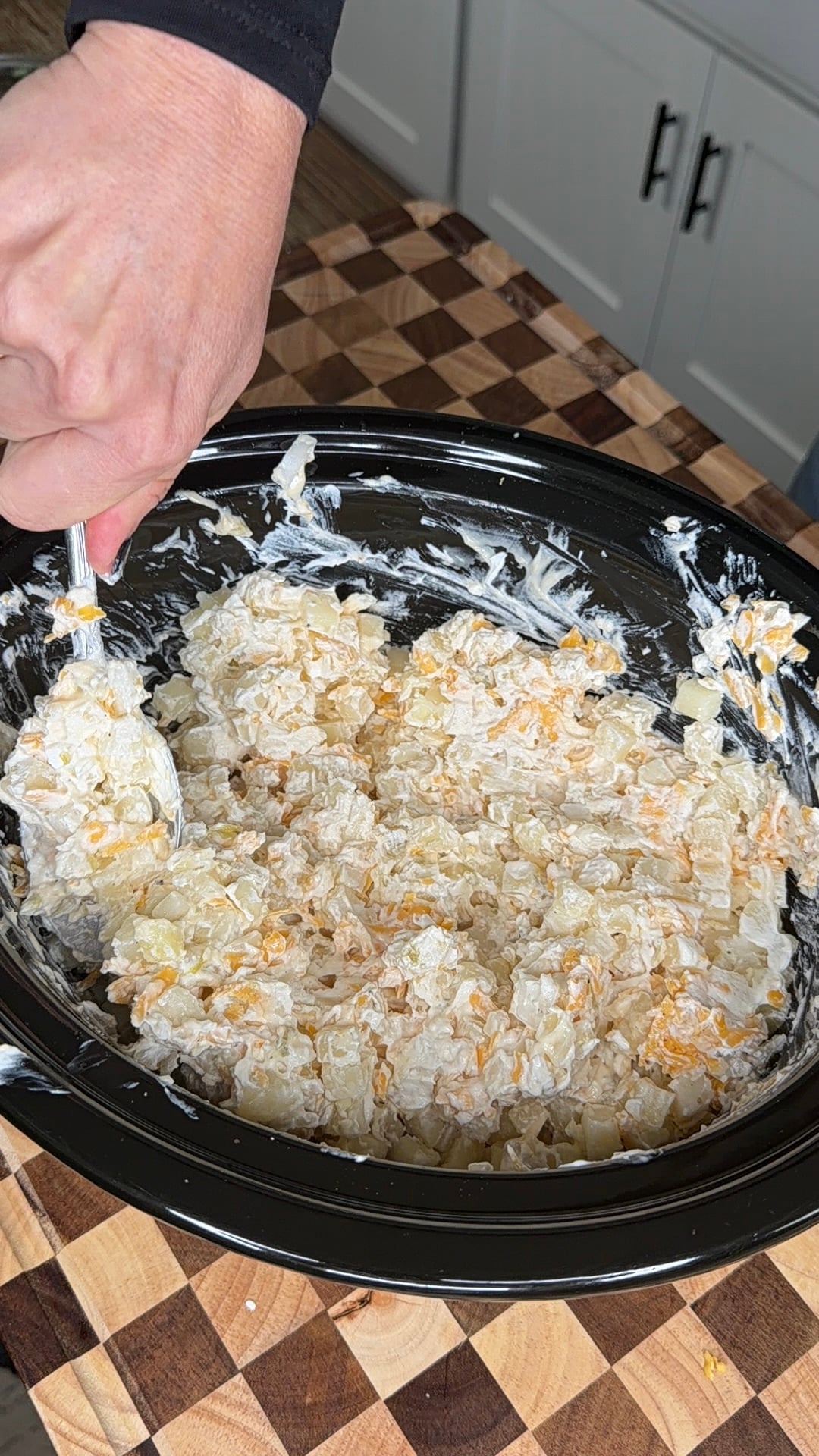 A person uses a spoon to stir a creamy mixture of cubed potatoes and shredded cheese inside a black slow cooker.