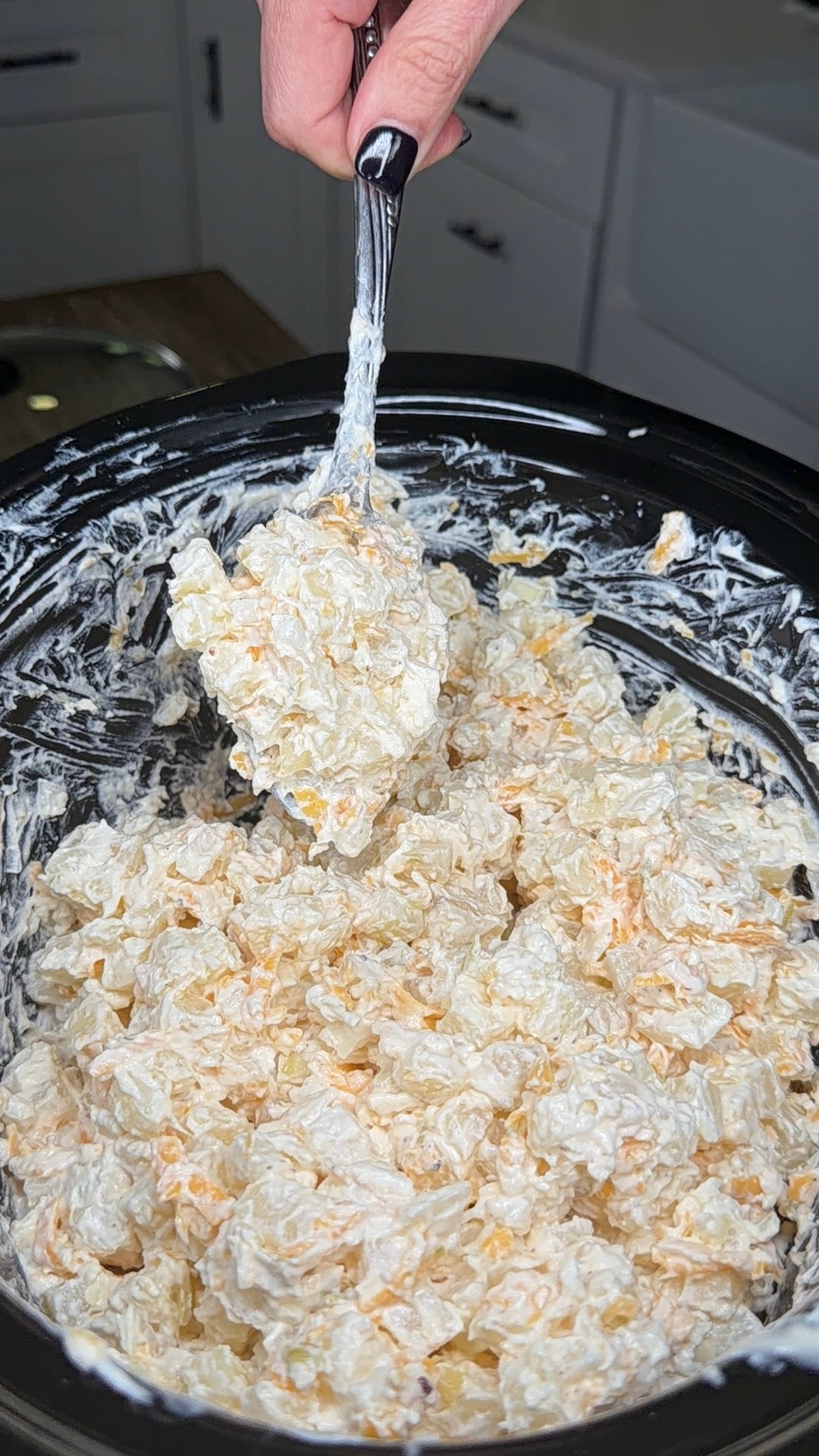 A hand holding a large metal spoon lifts a scoop of creamy potato salad with shredded cheese from a slow cooker.