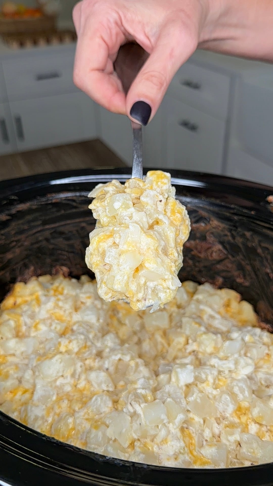 A hand holding a spoon over a slow cooker filled with creamy cheesy potatoes.