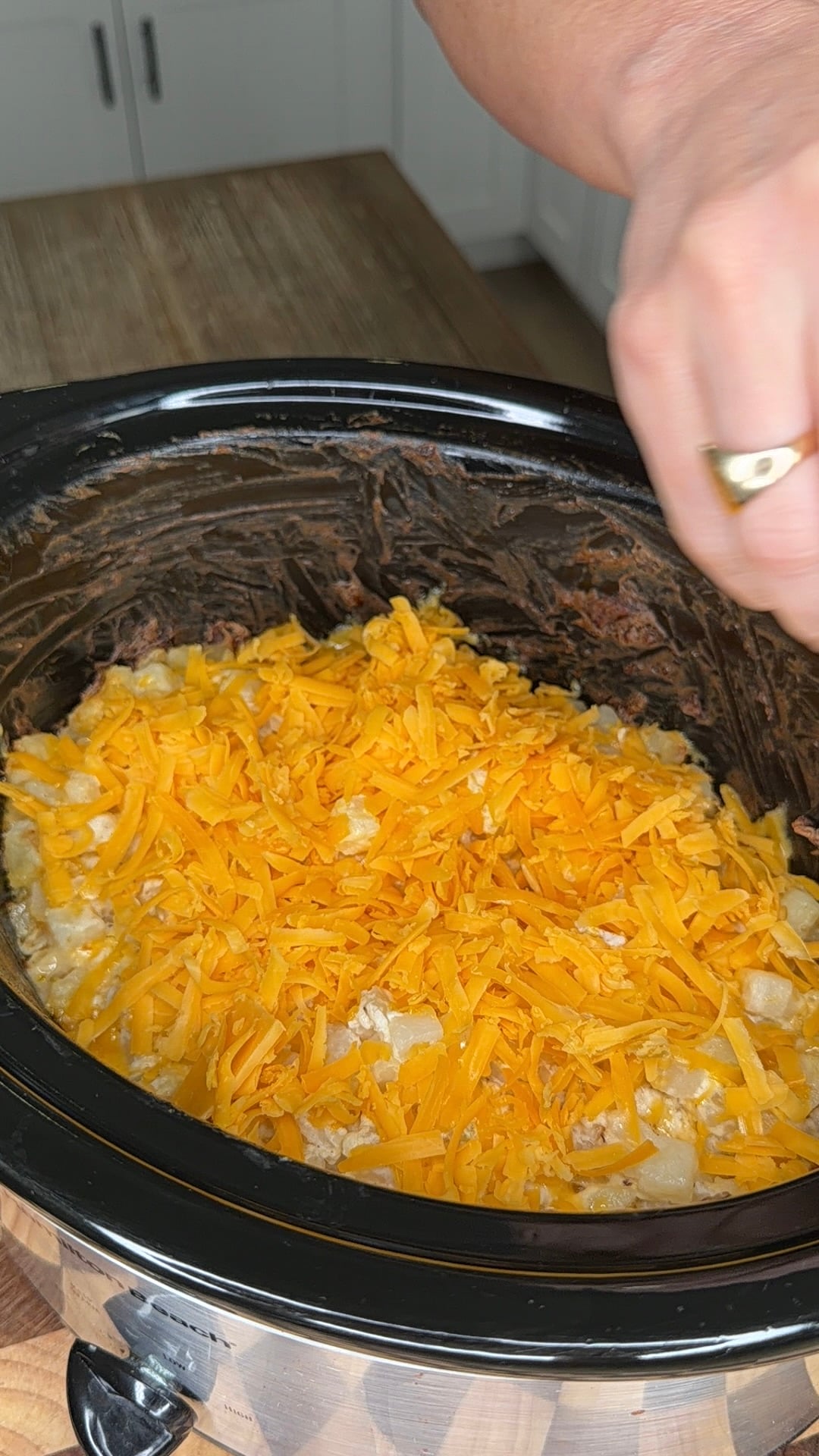 A hand sprinkles shredded cheddar cheese over a mixture in a slow cooker.