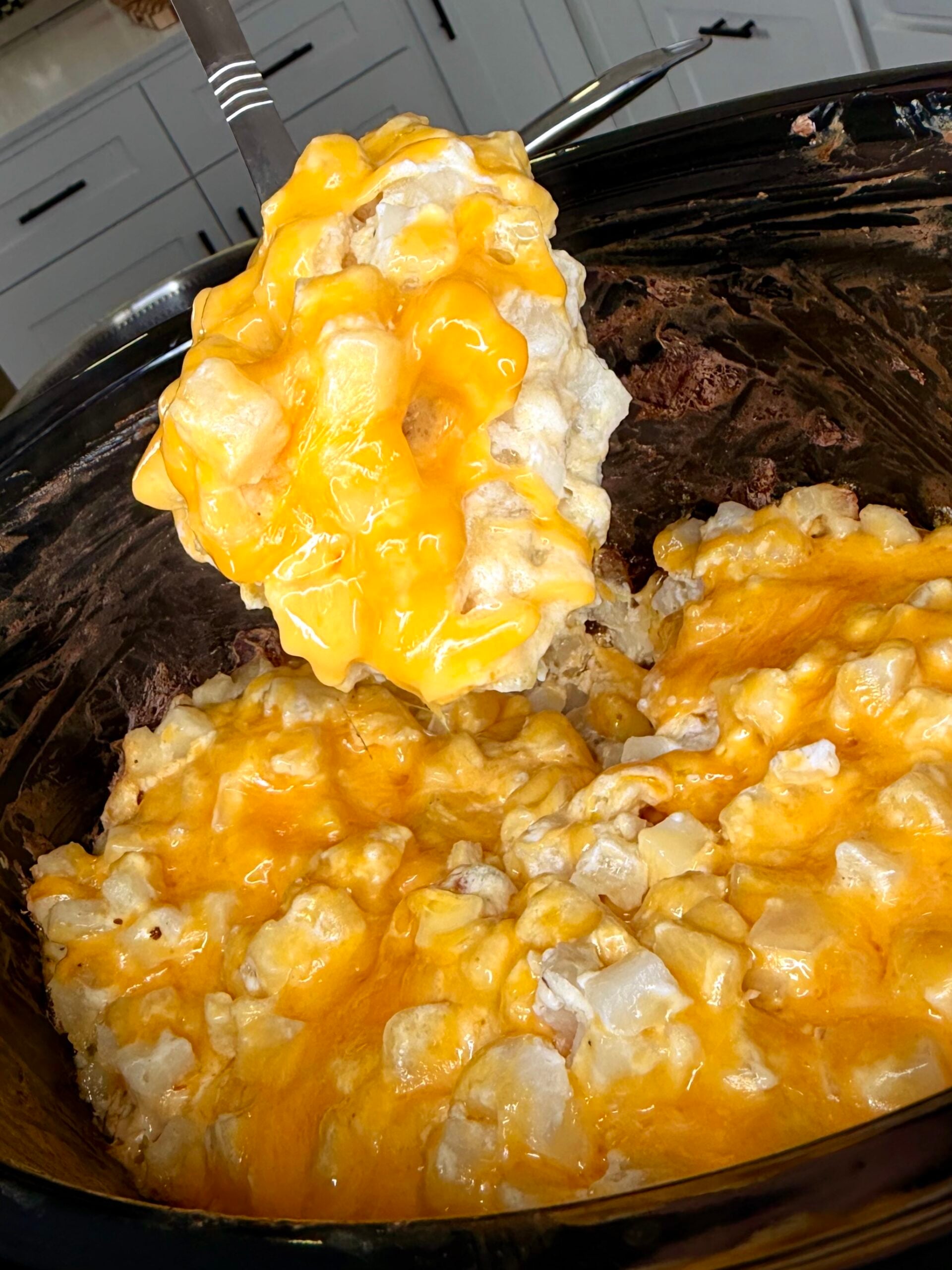 A metal spoon lifts a serving of cheesy potato casserole from a slow cooker.