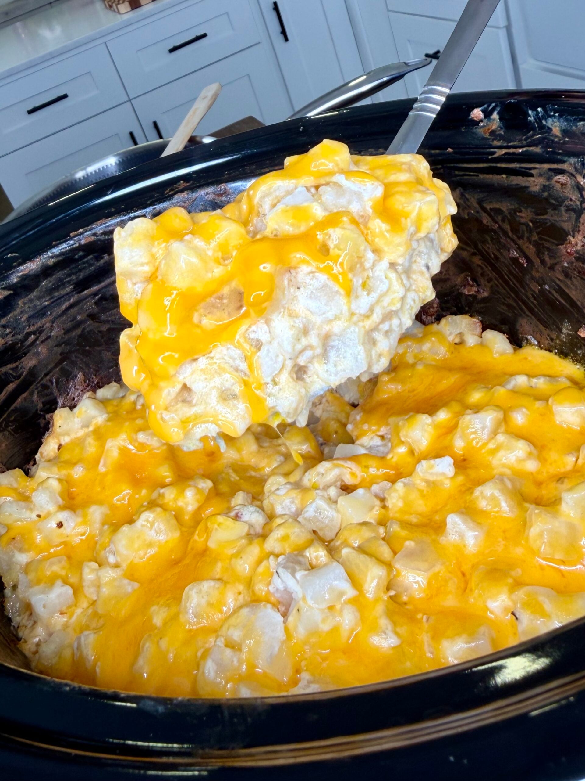 A metal spoon lifting a serving of cheesy potatoes from a black slow cooker.