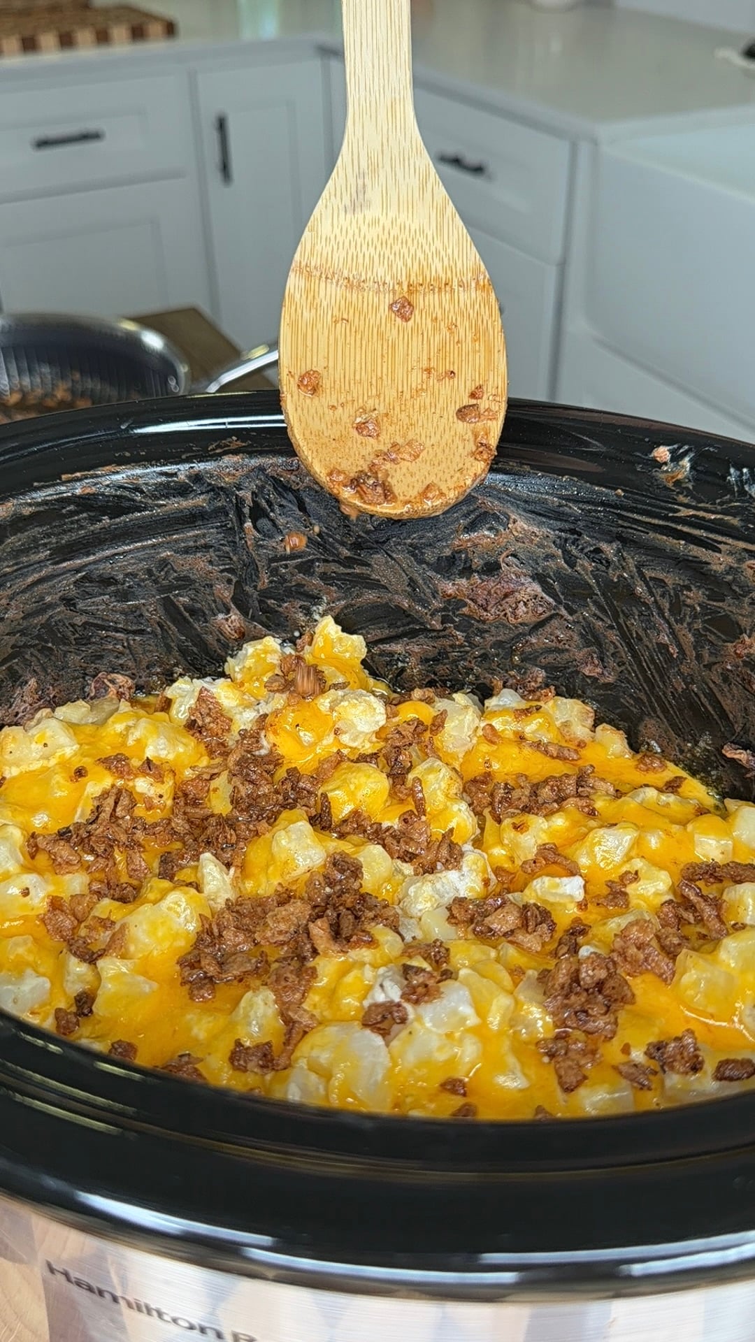 A wooden spoon held over a slow cooker filled with cheesy potatoes topped with bacon bits.
