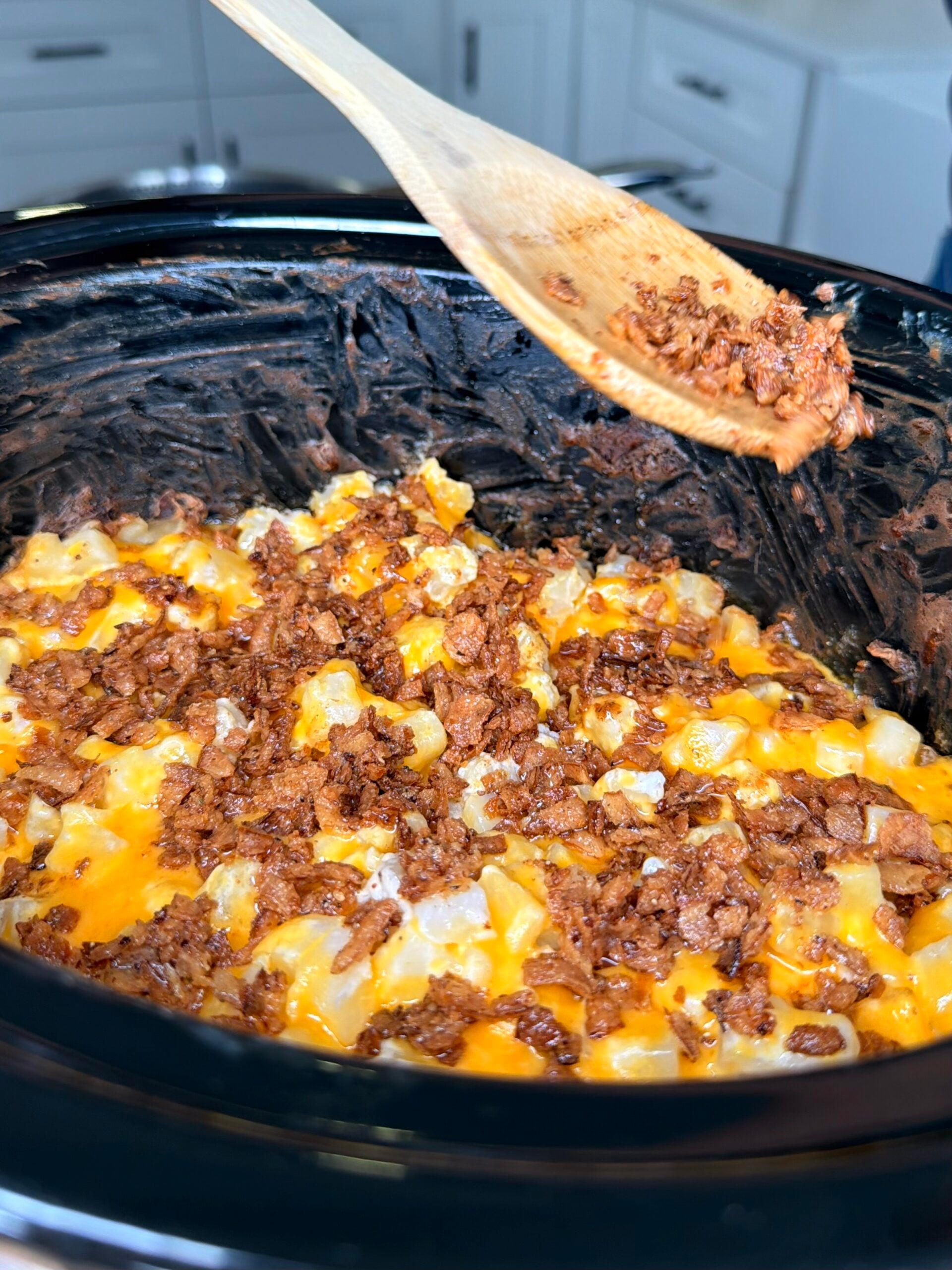 A slow cooker filled with cheesy potatoes topped with bacon crumbles, with a wooden spoon holding a scoop of bacon above.