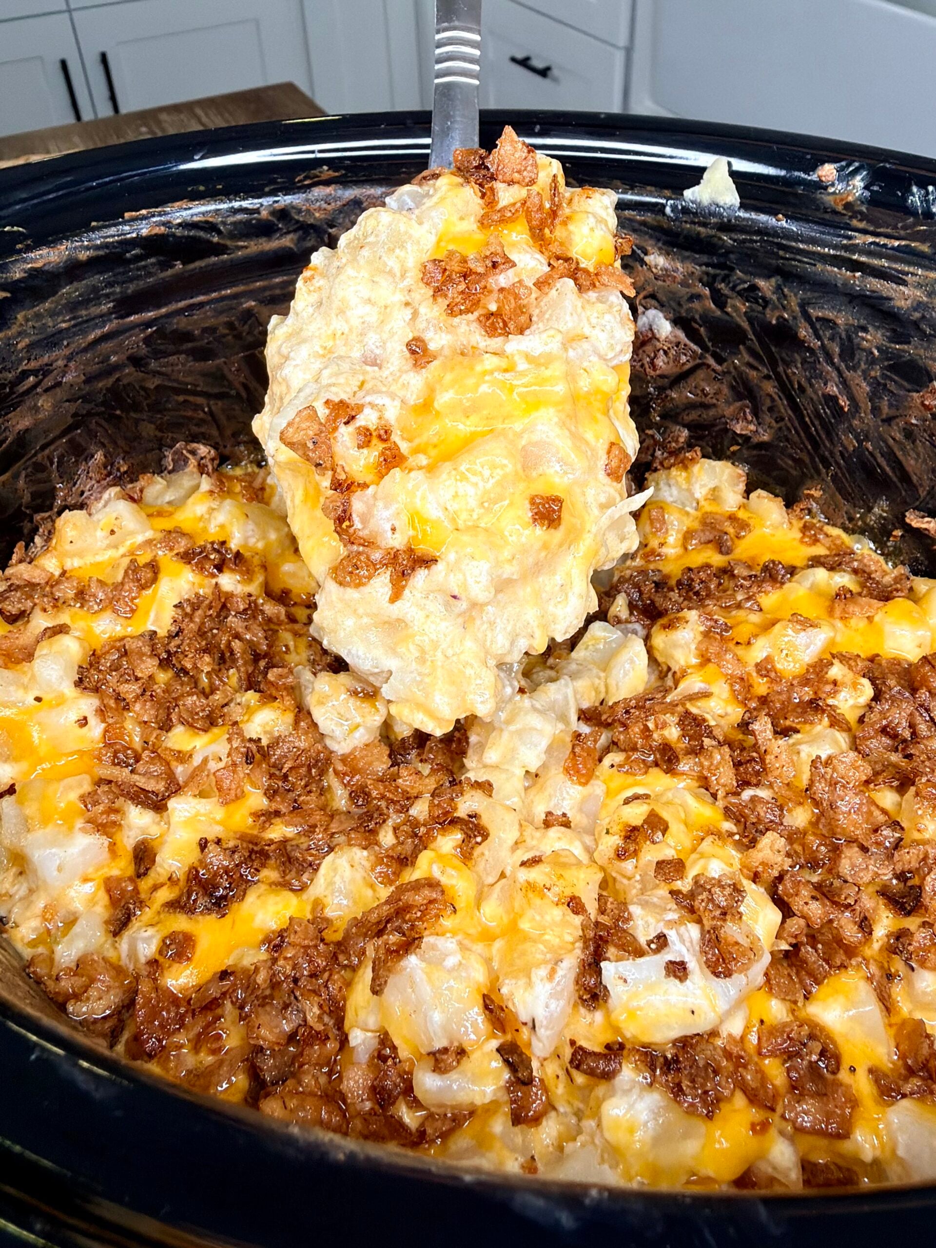 A serving spoon lifts a scoop of cheesy potato casserole topped with crispy bacon bits from a slow cooker.