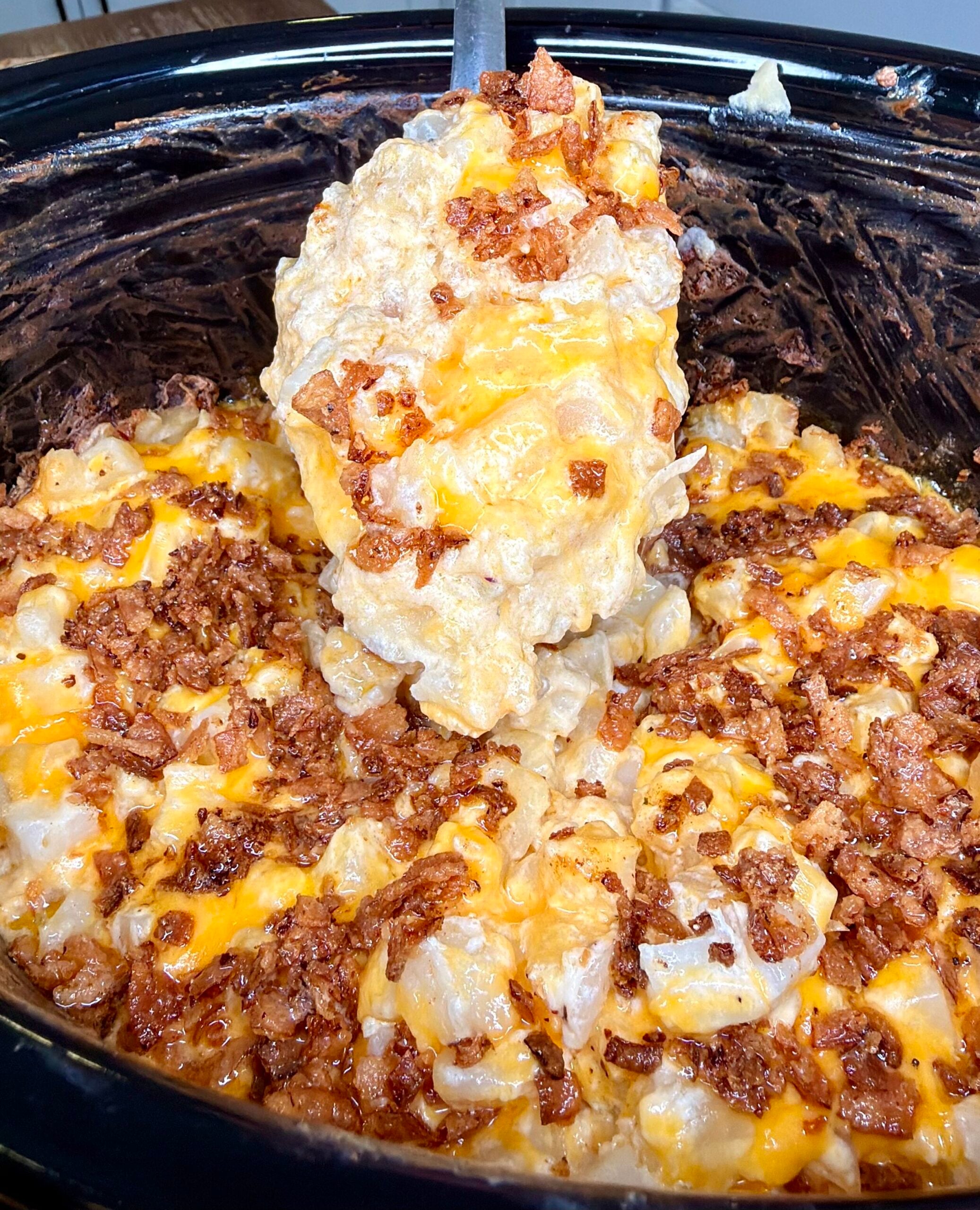 A metal spoon lifting a serving of cheesy potato casserole topped with bacon bits from a slow cooker.
