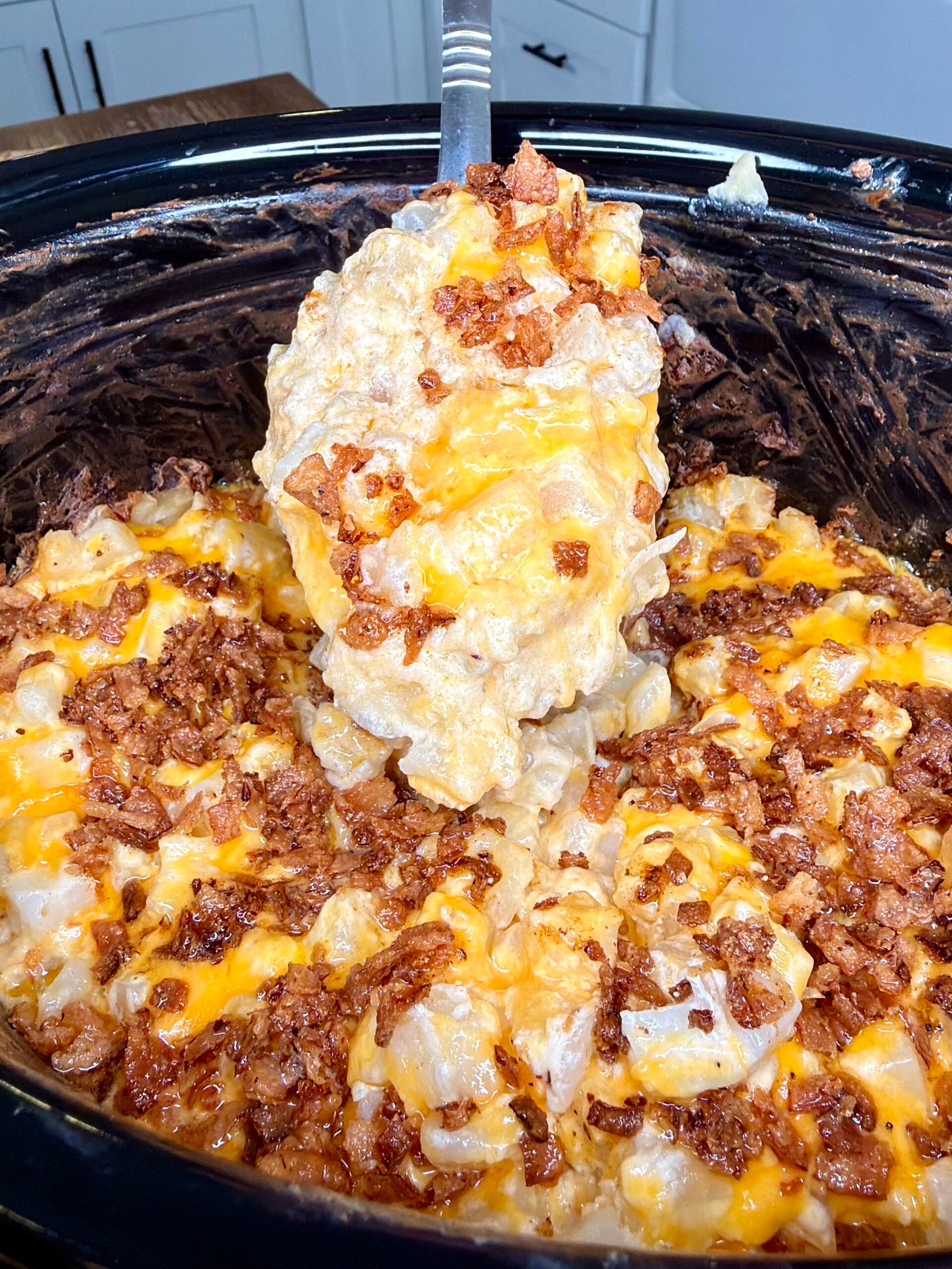 A metal spoon lifting a serving of cheesy potato casserole topped with bacon bits from a slow cooker.