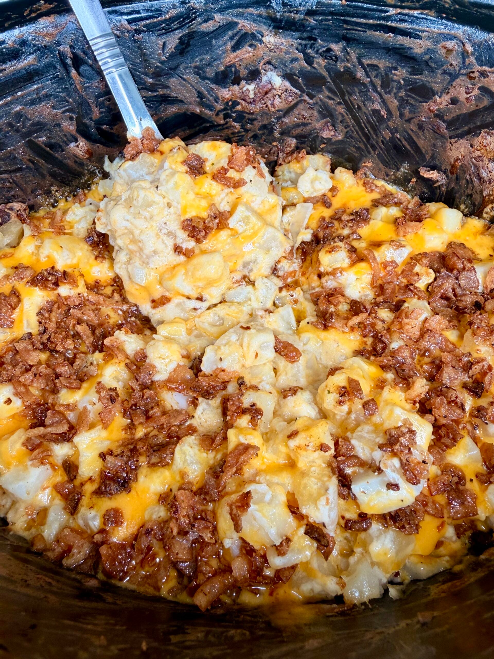 A slow cooker filled with cheesy potatoes topped with bacon bits, with a metal serving spoon lifting a portion.