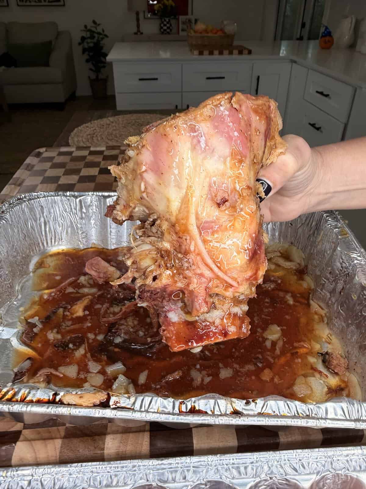 A hand holds a large, cooked pork rib over a disposable aluminum roasting pan filled with sauce and onions.